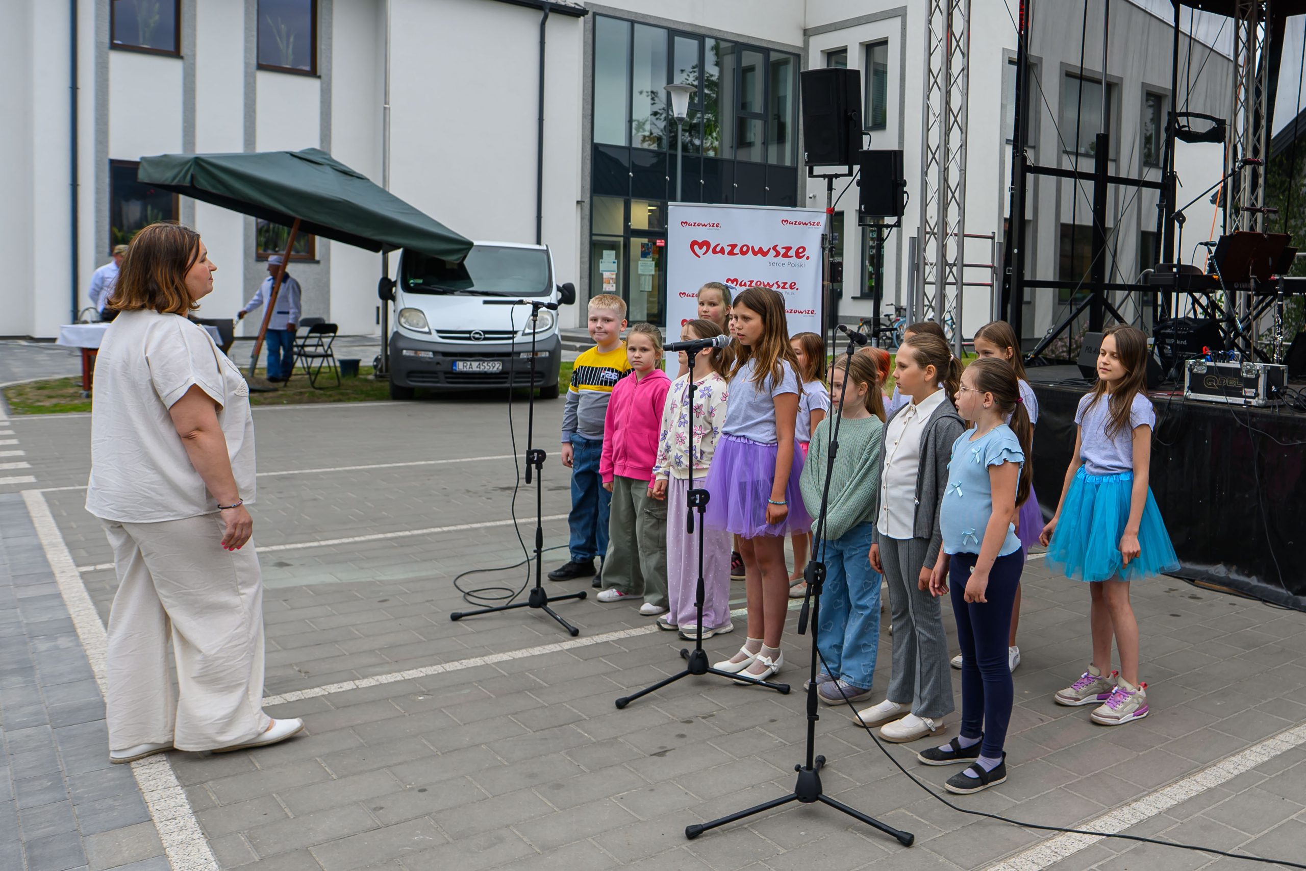 Grupa dzieci w kolorowych ubraniach śpiewa na scenie z mikrofonami. Przed nimi stoi kobieta. W tle biały budynek i samochód.