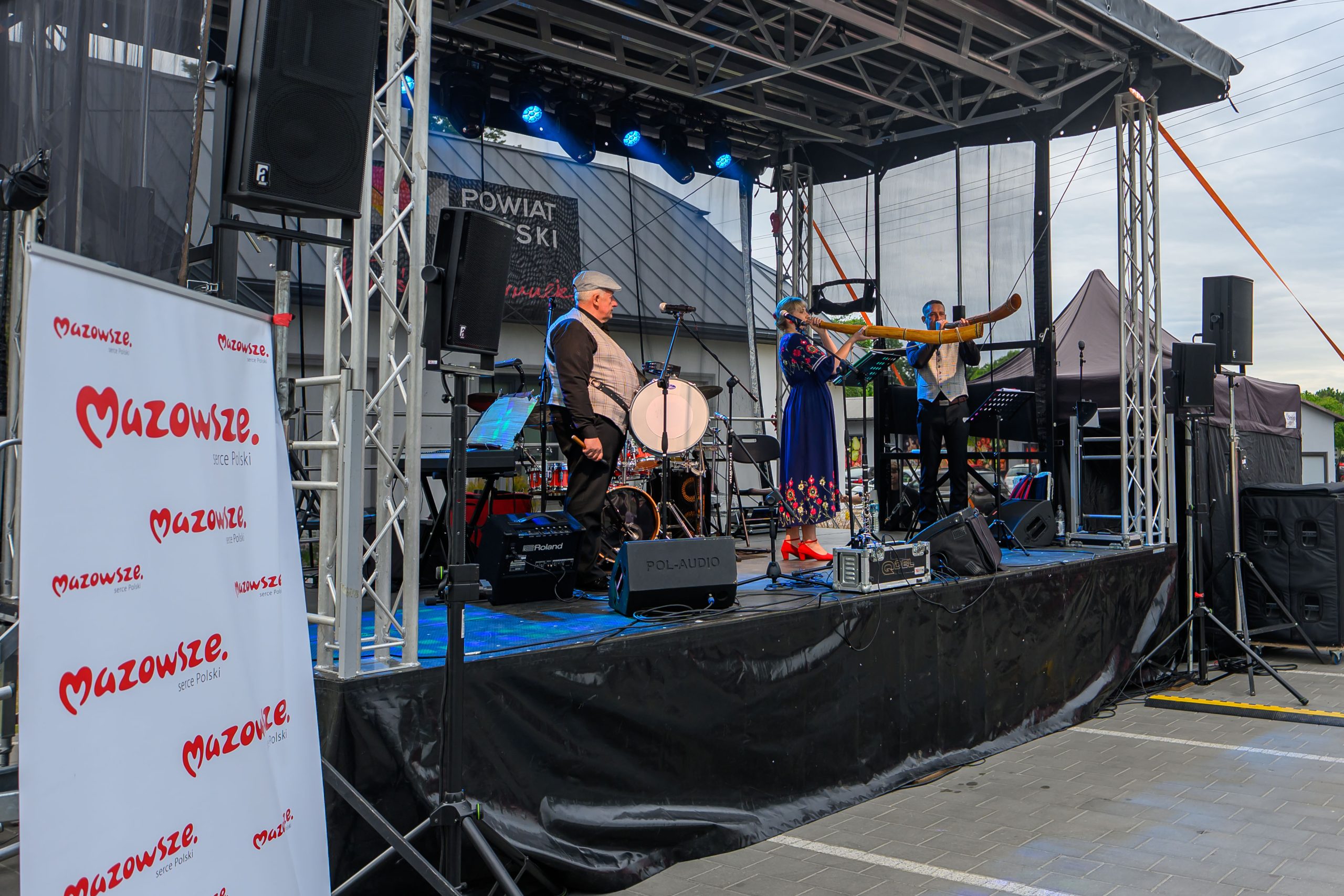 Zespół muzyczny na scenie na tle budynku, gra na instrumentach. Dominują kolory niebieski, czarny i czerwony. Baner z napisem Mazowsze.