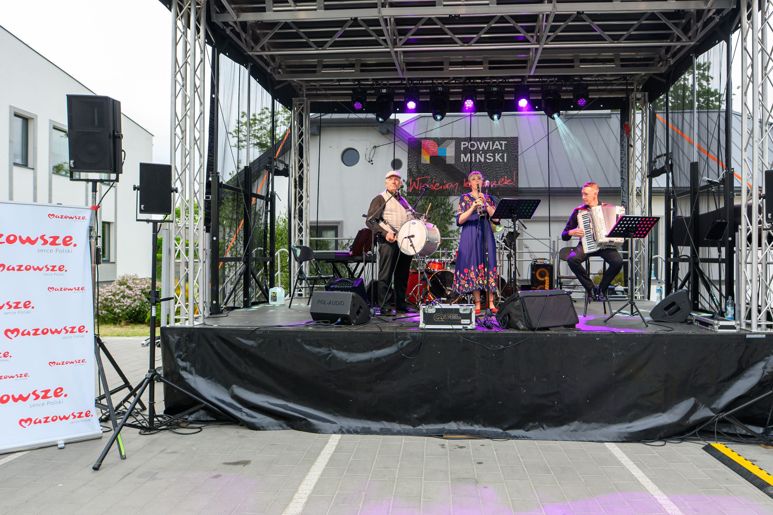 Trzech muzyków gra na scenie: kobieta śpiewa, mężczyzna gra na akordeonie, trzeci na bębnie. W tle banner Powiat Miński. Tło jasne, szaro-białe budynki.