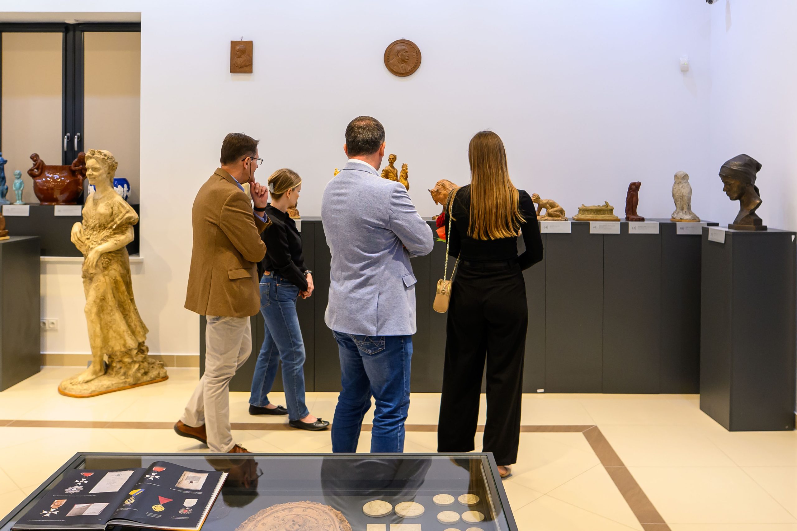 Grupa osób oglądająca wystawę rzeźb w jasnej sali muzeum. Dominują kolory beżowy i szary. W tle na ścianie wiszą małe płaskorzeźby.