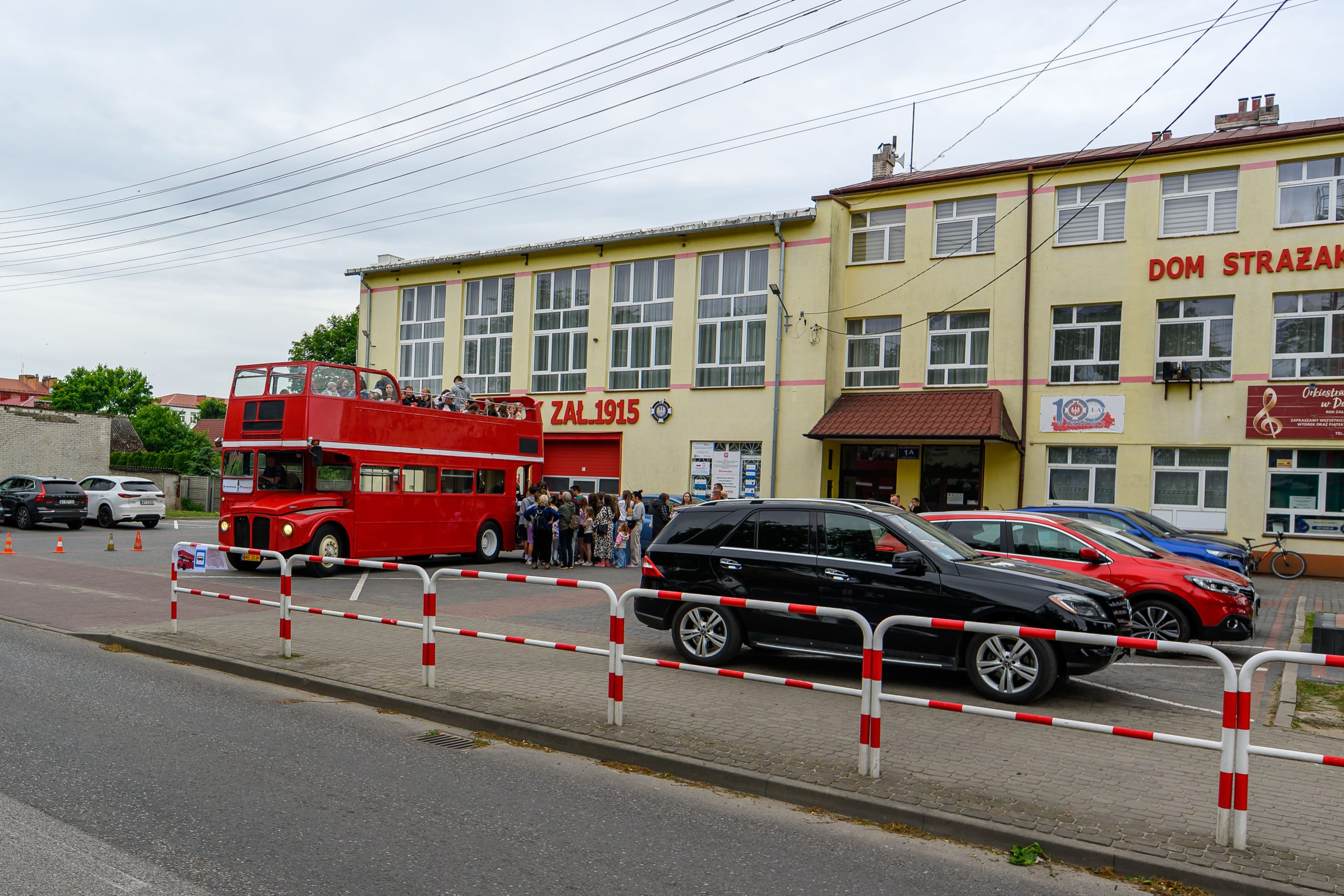 Czerwony, piętrowy autobus stoi przed żółtym budynkiem z napisem Dom Strażaka. Na ulicy kilka zaparkowanych samochodów. Grupa ludzi stoi obok autobusu.