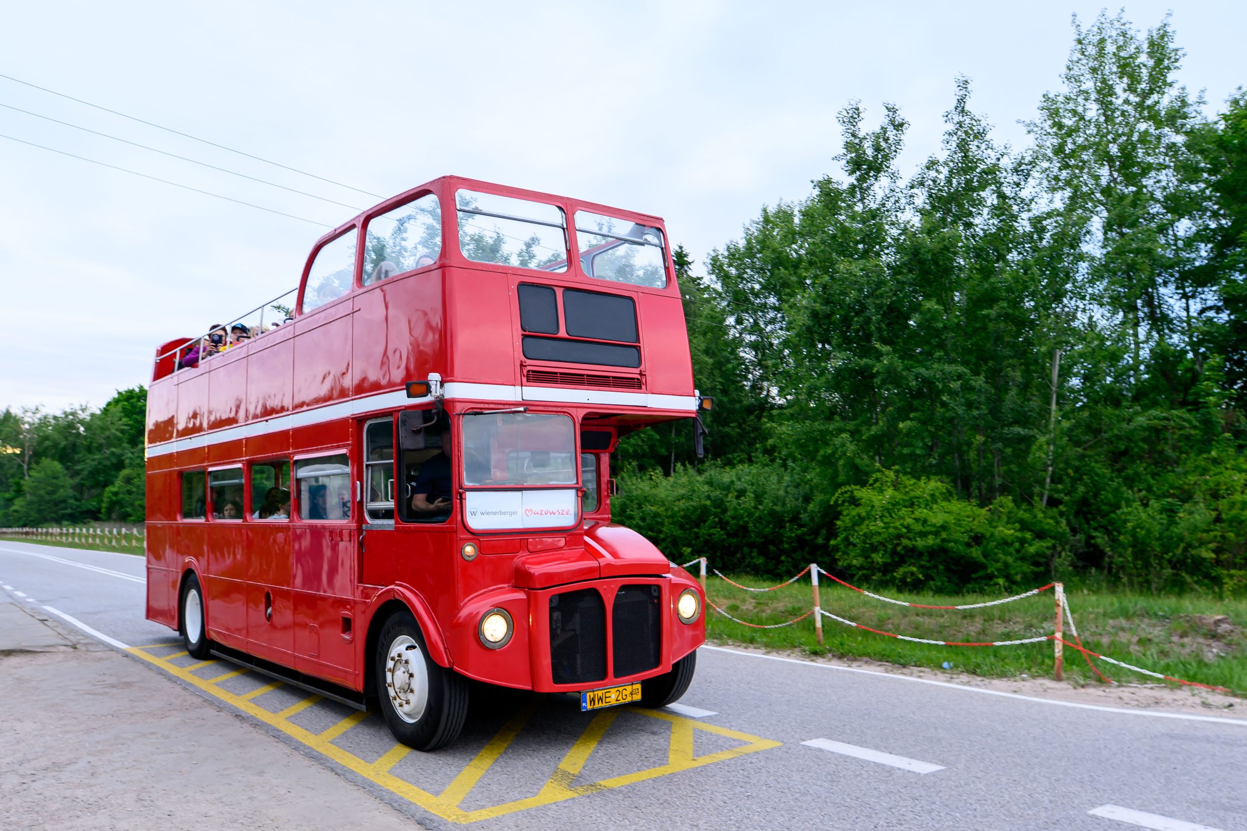 Czerwony piętrowy autobus jedzie po drodze. W tle zielone drzewa. Niebo lekko zachmurzone.