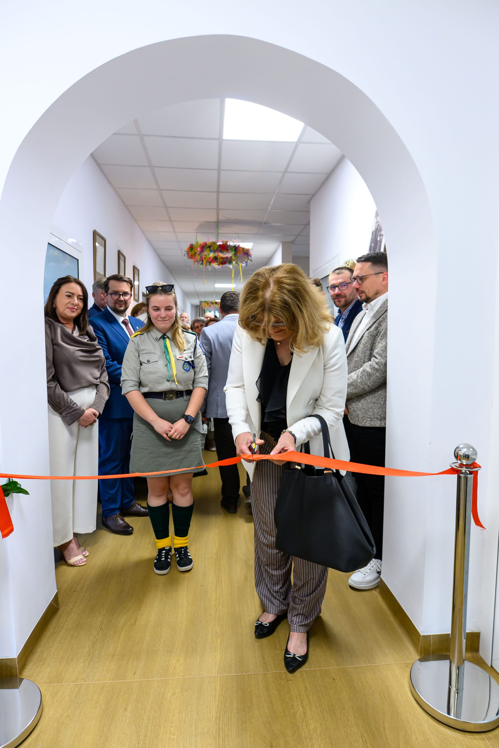 Funkcjonariuszka przecina wstęgę w jasnym korytarzu, otoczona ludźmi. Kobieta w białej marynarce i wzorzystych spodniach. Drewniana podłoga, ściany białe z zawieszonymi dekoracjami.