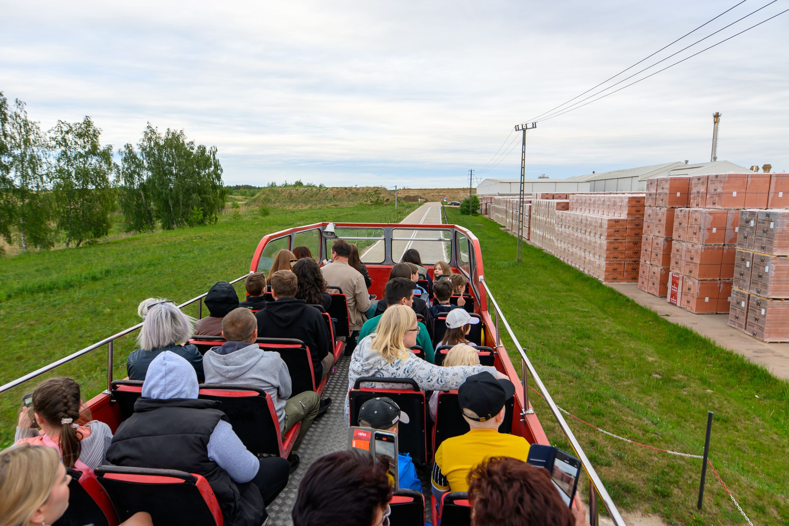 Ludzie siedzą na otwartym piętrze czerwonego autobusu i patrzą na fabrykę cegieł. Po lewej stronie zielona trawa i drzewa, po prawej stosy cegieł. Niebo jest pochmurne.