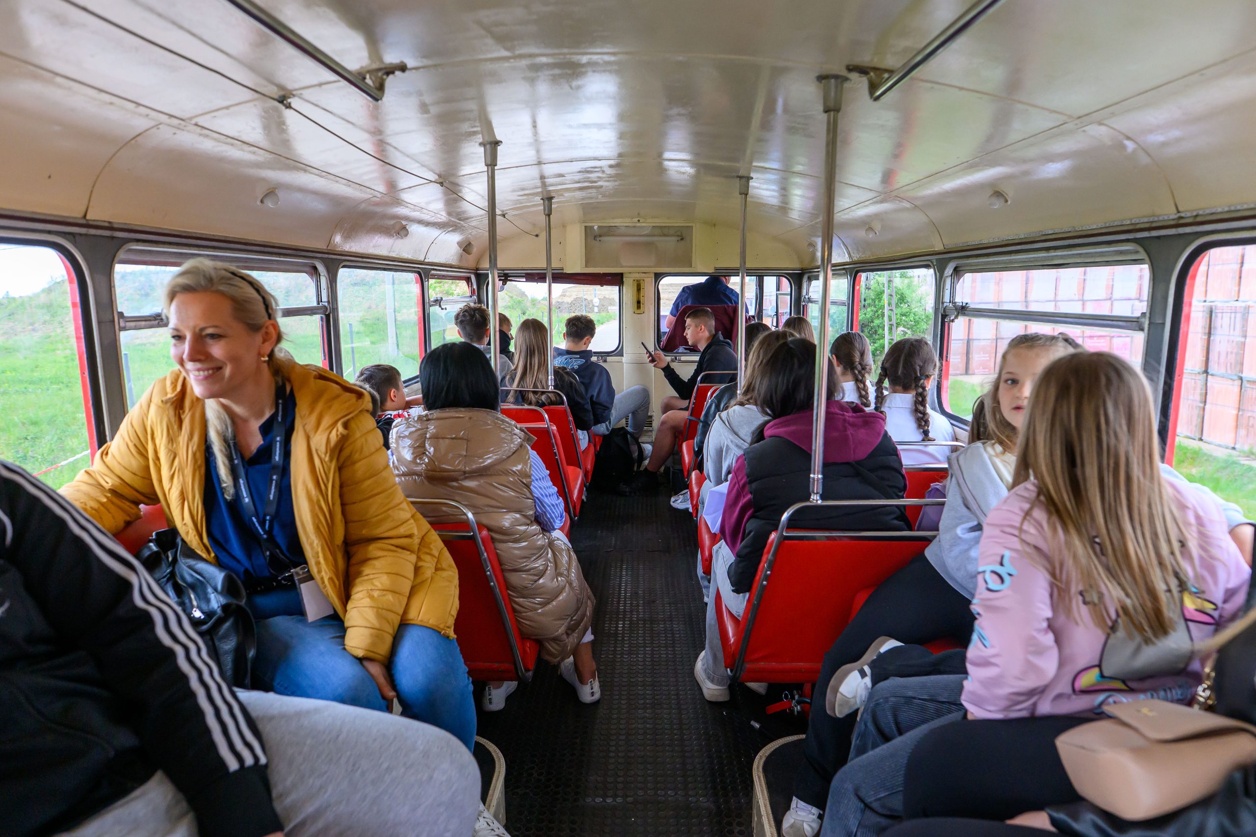 Ludzie siedzą w starym autobusie z czerwonymi siedzeniami. Tło jest zielone z widokiem przez okna. Kobieta w żółtej kurtce uśmiecha się.