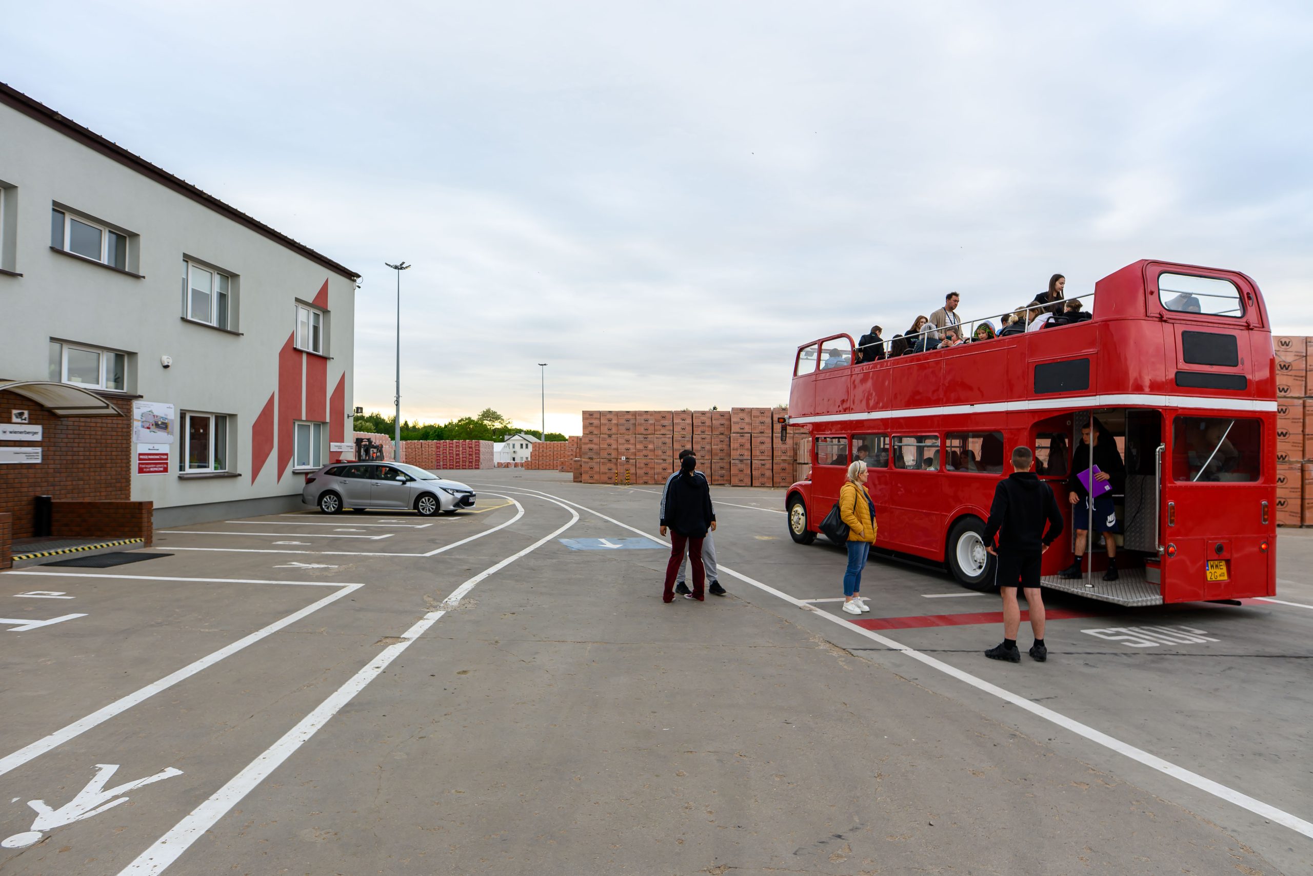 Czerwony, dwupoziomowy autobus stoi na placu z ludźmi na górnym pokładzie. Szary budynek po lewej i stosy cegieł w tle. Pochmurny dzień.