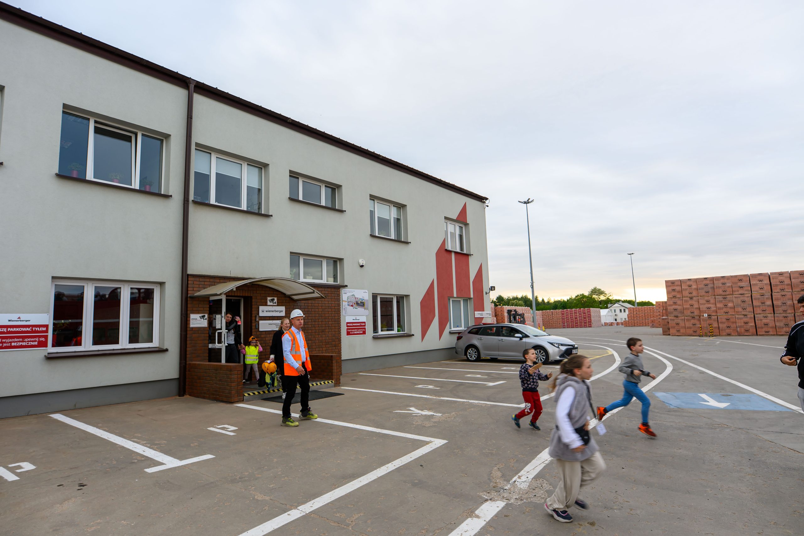 Na zdjęciu znajduje się szara, dwupiętrowa budynek z czerwonymi elementami. Przed nim ludzie, w tym dzieci biegające po parkingu. W tle widać stosy cegieł i fragmenty zieleni.