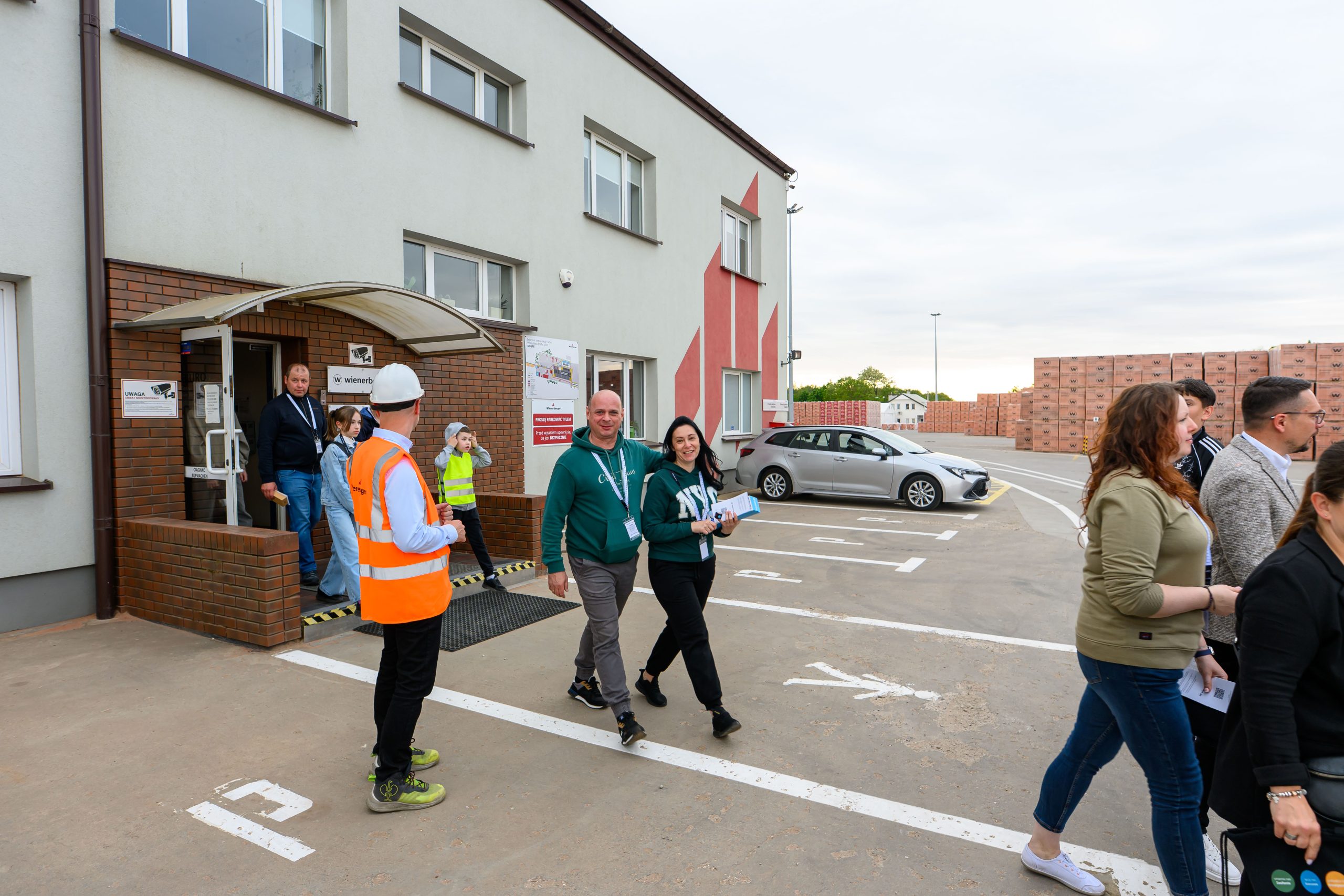 Grupa ludzi wychodzi z budynku w beżowo-czerwonym kolorze. Mężczyzna w pomarańczowej kamizelce stoi na zewnątrz. W tle, parking i stosy cegieł.