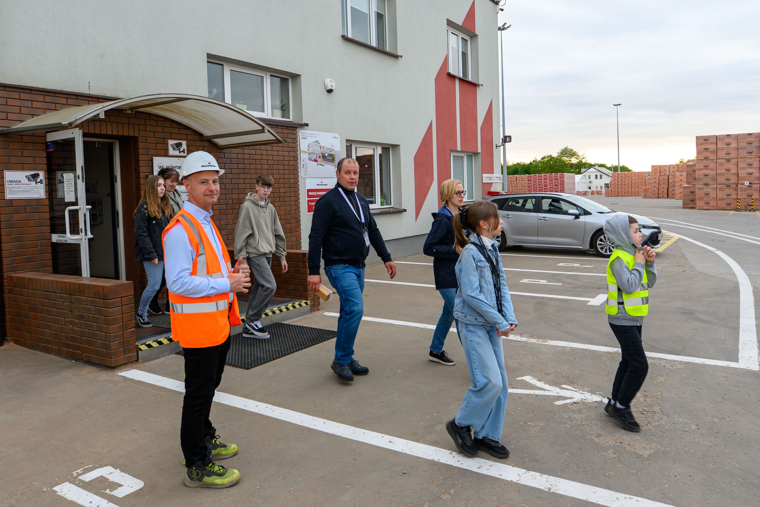 Grupa ludzi w kamizelkach odblaskowych wychodzi z budynku z cegły. Na parkingu widać samochód i stosy kartonów. Tło jest zachmurzone.