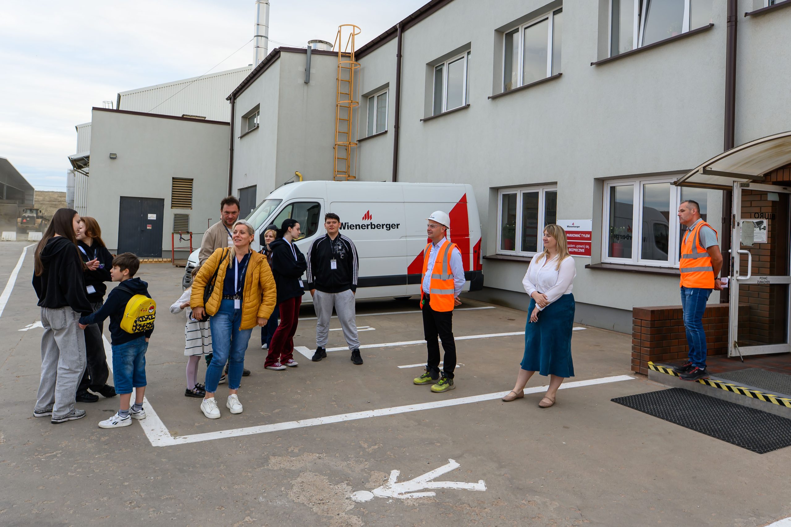 Grupa osób przed zakładem przemysłowym, z białym vanem w tle. Jasne budynki, niektóre osoby w kamizelkach odblaskowych. Tło jest szare i przemysłowe.