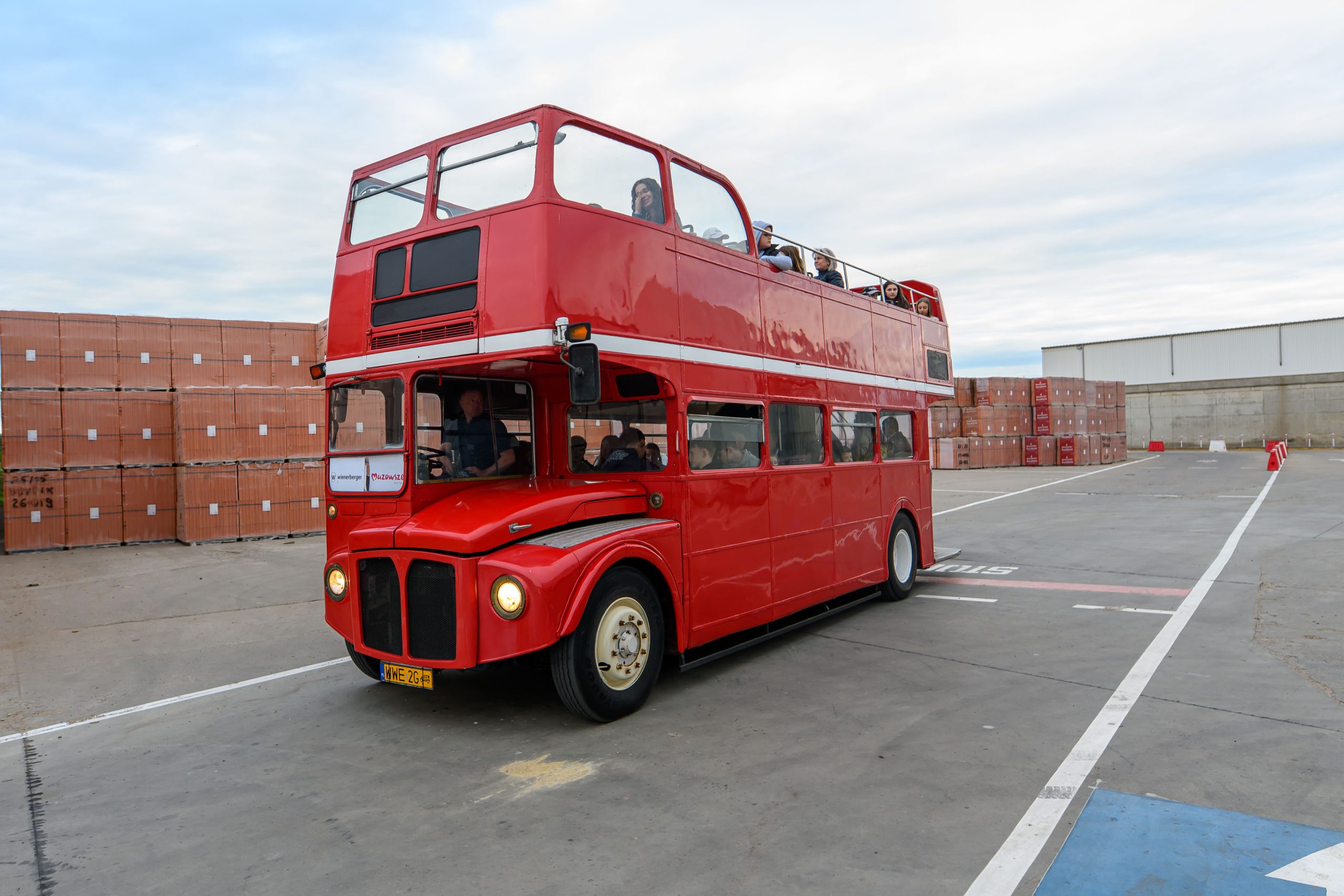 Czerwony piętrowy autobus jedzie po betonowej powierzchni placu. W tle stosy kartonowych pudeł i budynek. Na górnym pokładzie siedzą pasażerowie.