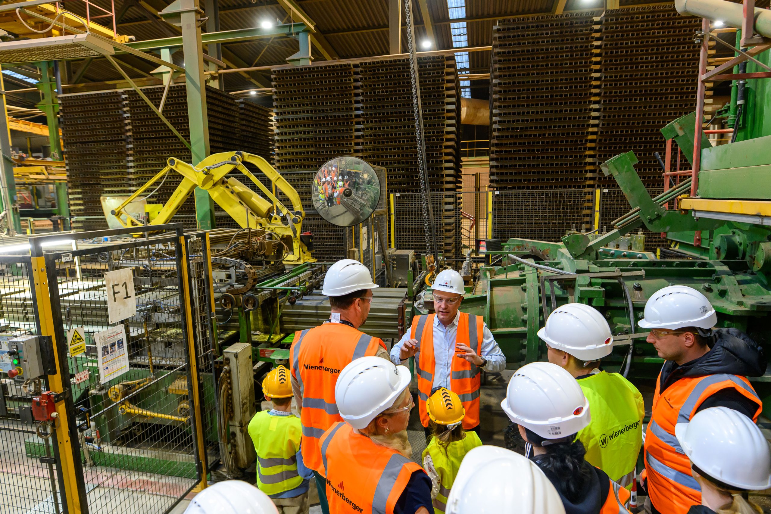 Grupa osób w hełmach i pomarańczowych kamizelkach jest w fabryce. Tło to maszyny i duże metalowe stosy. Przewodnik wyjaśnia procesy produkcyjne.