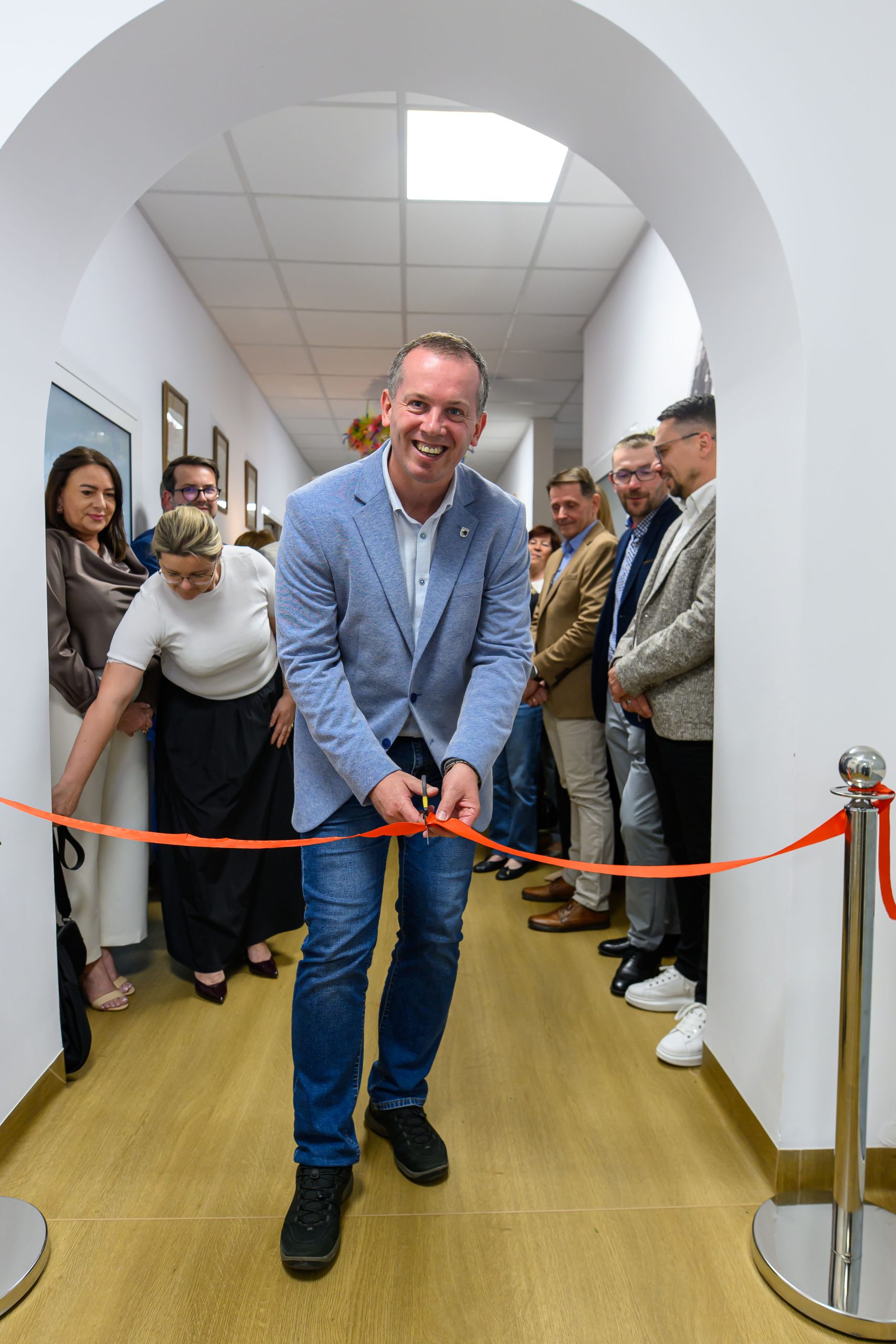 Mężczyzna w niebieskiej marynarce przecina czerwoną wstęgę podczas ceremonii otwarcia. W tle grupa uśmiechniętych osób na korytarzu. Dominują kolory bieli i drewna.
