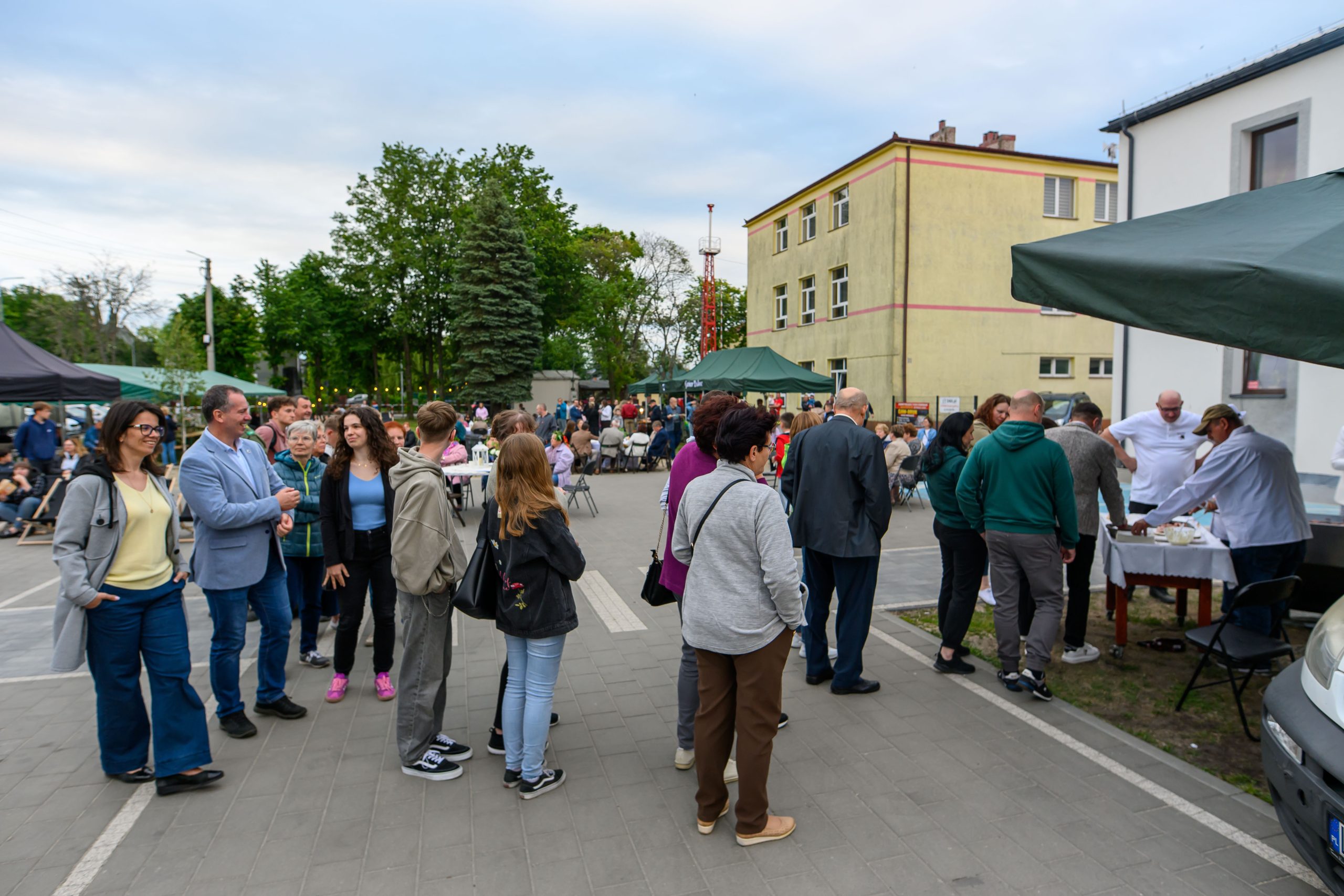Grupa ludzi na placu przed budynkami, zielone i beżowe namioty. Drzewa w tle, niebo jasne, ludzie rozmawiają.