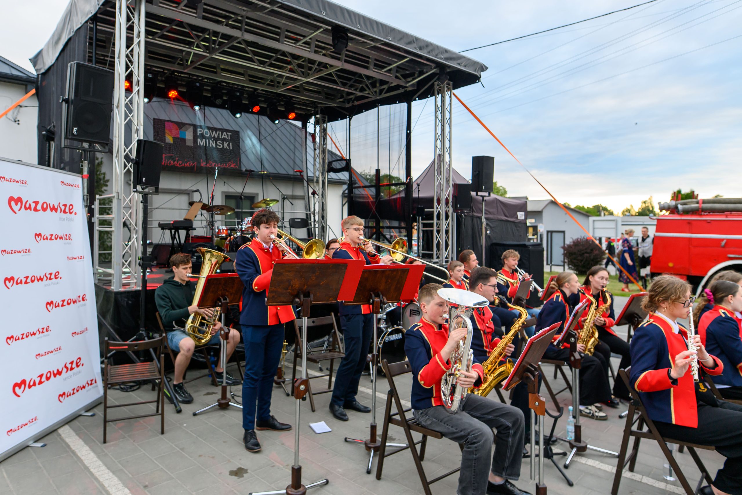 Orkiestra na scenie, młodzież w granatowych uniformach z czerwonymi elementami. Tło stanowi namiot z napisem Powiat Miński. Obok scena z instrumentami i mikrofonami.