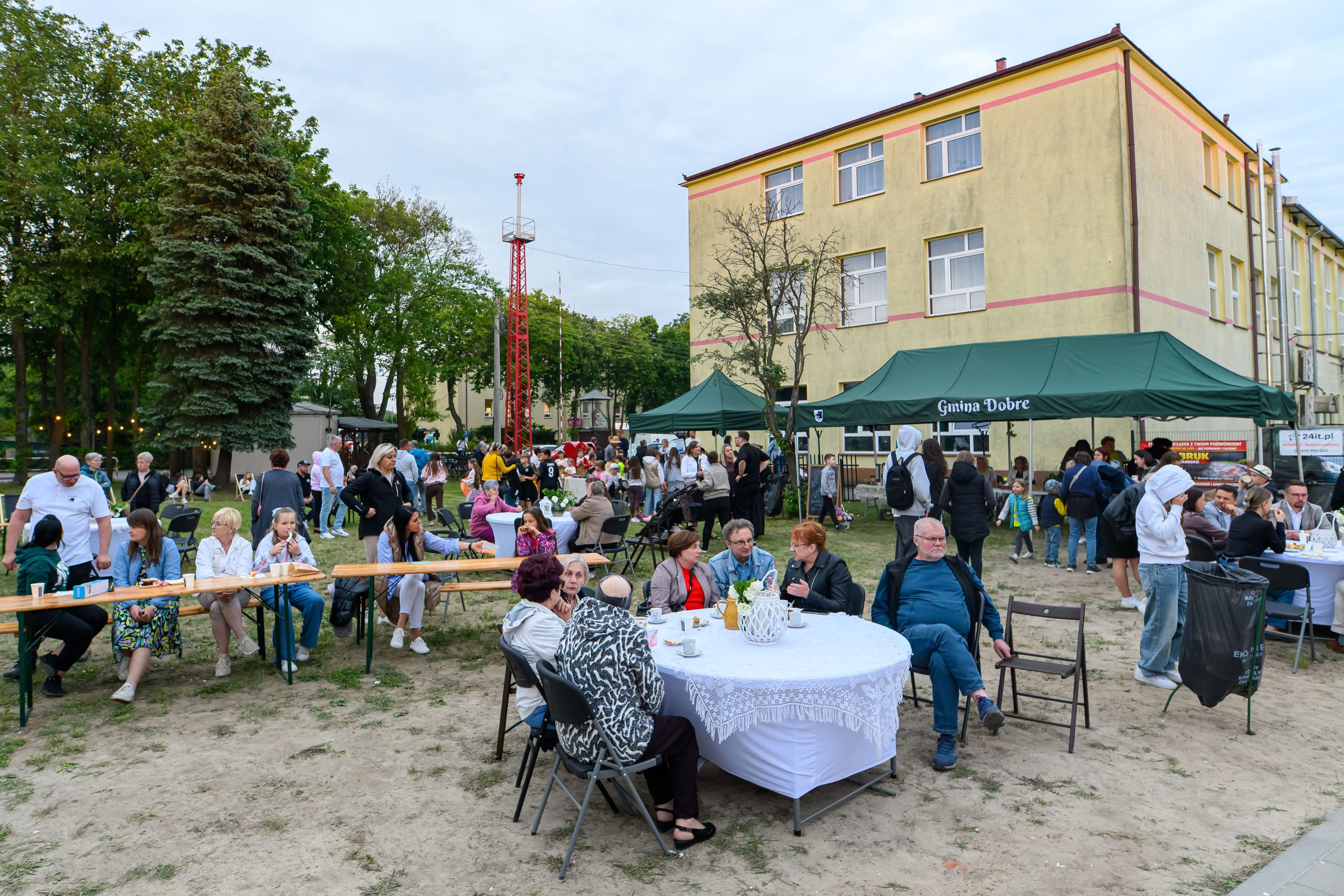 Na zdjęciu widać grupę ludzi na pikniku, siedzących przy stołach na zewnątrz. W tle jest zielony namiot z napisem oraz żółty budynek. Trawa i drzewa otaczają miejsce wydarzenia, tworząc przyjemną atmosferę.