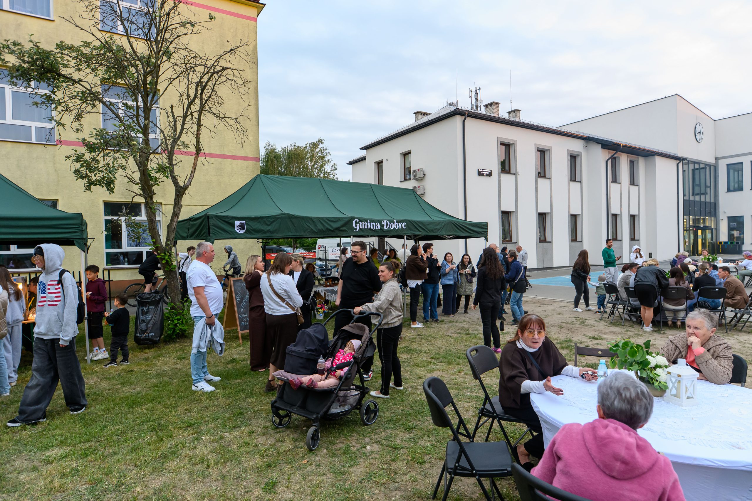 Grupa osób zgromadzona pod zielonym namiotem na tle budynku. Lato, na trawie stoliki z białymi obrusami. Swobodna, społeczna atmosfera.