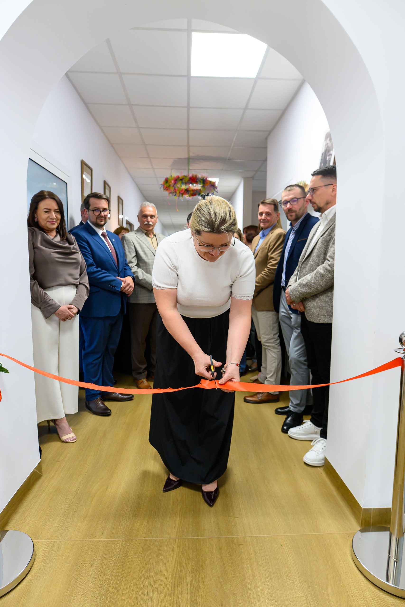 Kobieta przecina czerwoną wstęgę w korytarzu budynku. Tło jest białe z drewnianą podłogą. Obok stoi grupa elegancko ubranych osób.