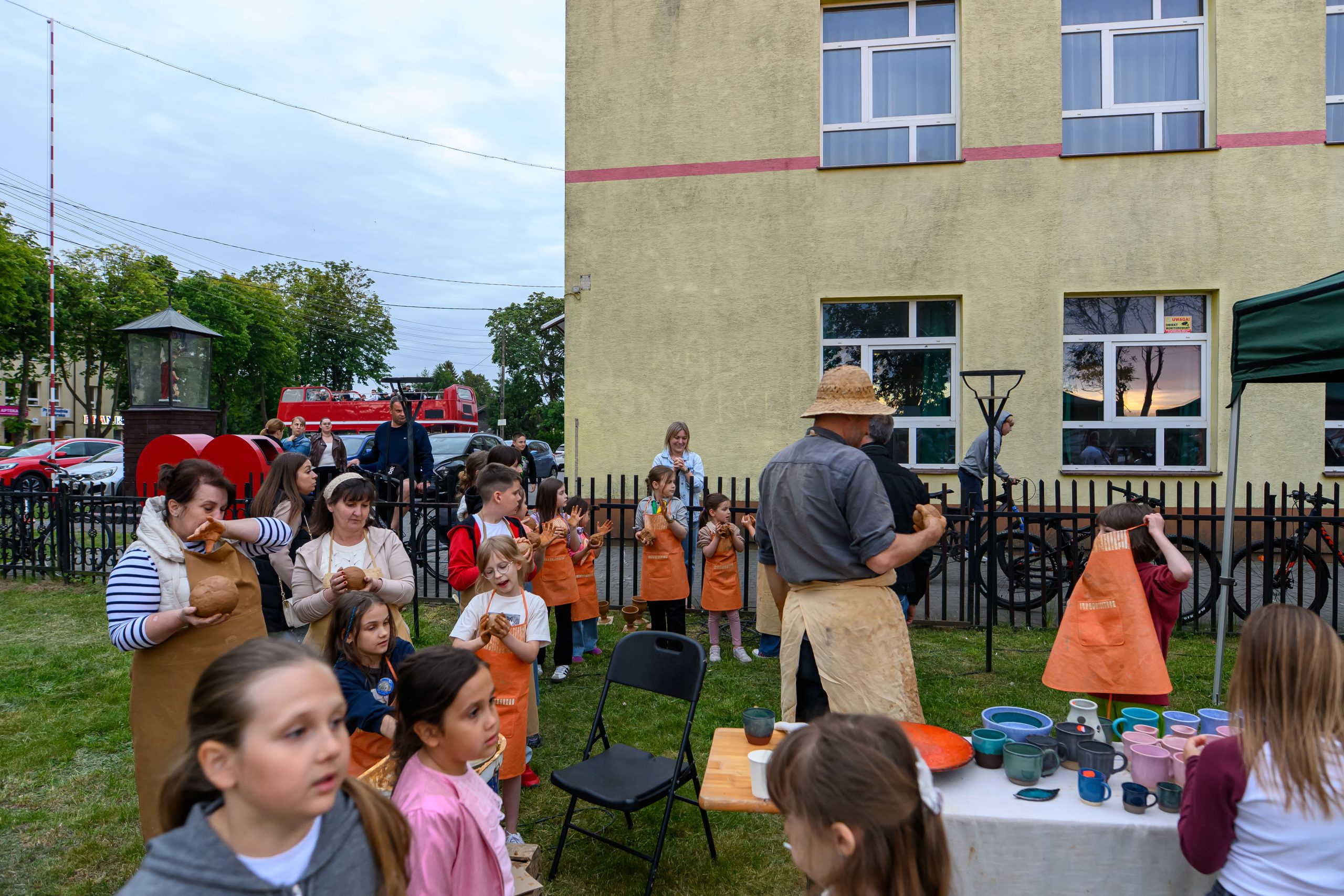 Grupa dzieci i dorosłych w fartuchach na warsztatach na świeżym powietrzu przy budynku. Stoły z kolorowymi naczyniami. W tle widać zielone drzewa i czerwony pojazd.