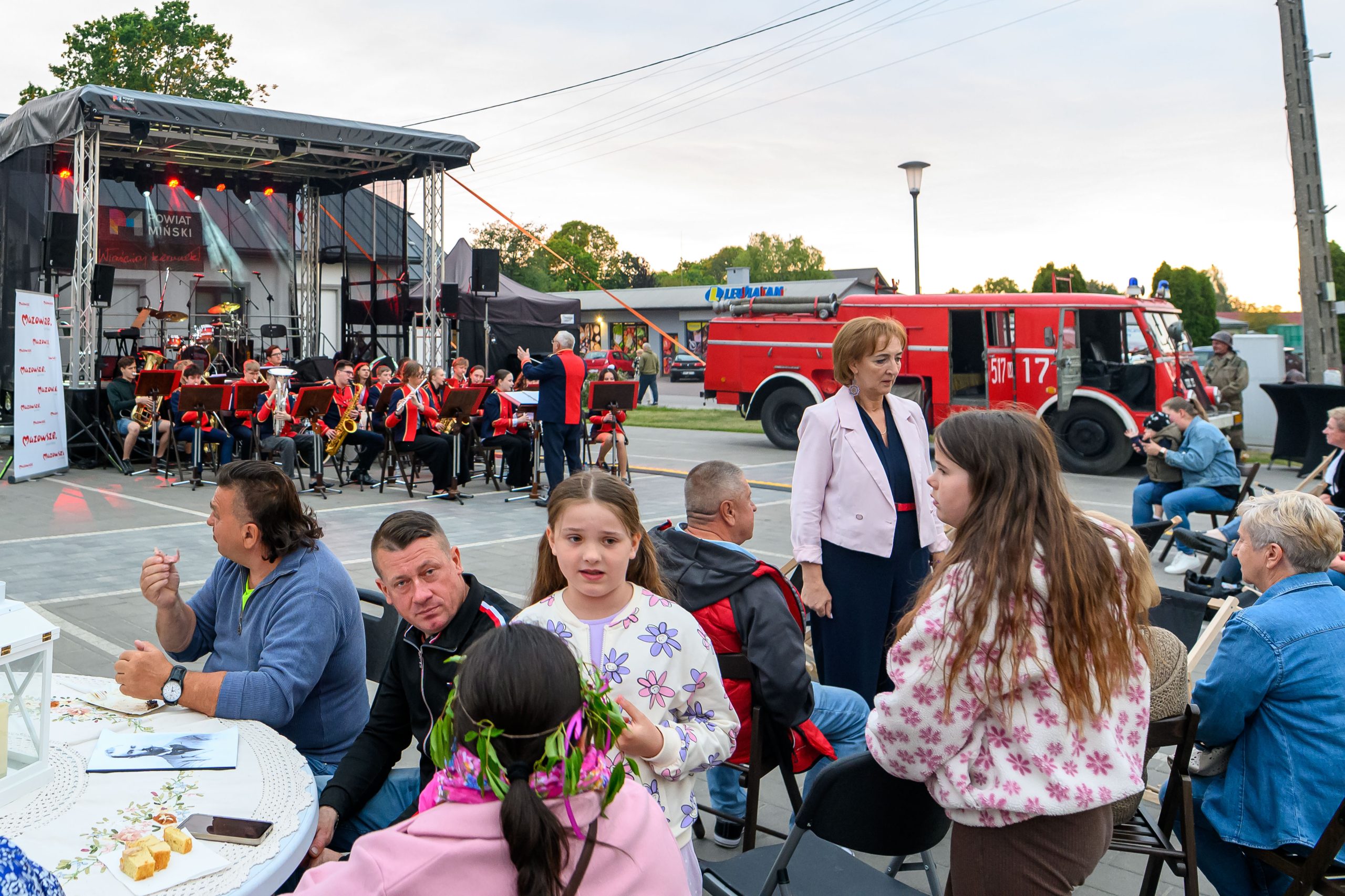 Na scenie orkiestra w czerwonych strojach przy instrumencie, w tle czerwony wóz strażacki. Przed sceną grupa ludzi siedzi i stoi, rozmawiając. Jest pogodny dzień, niebo lekko zachmurzone.
