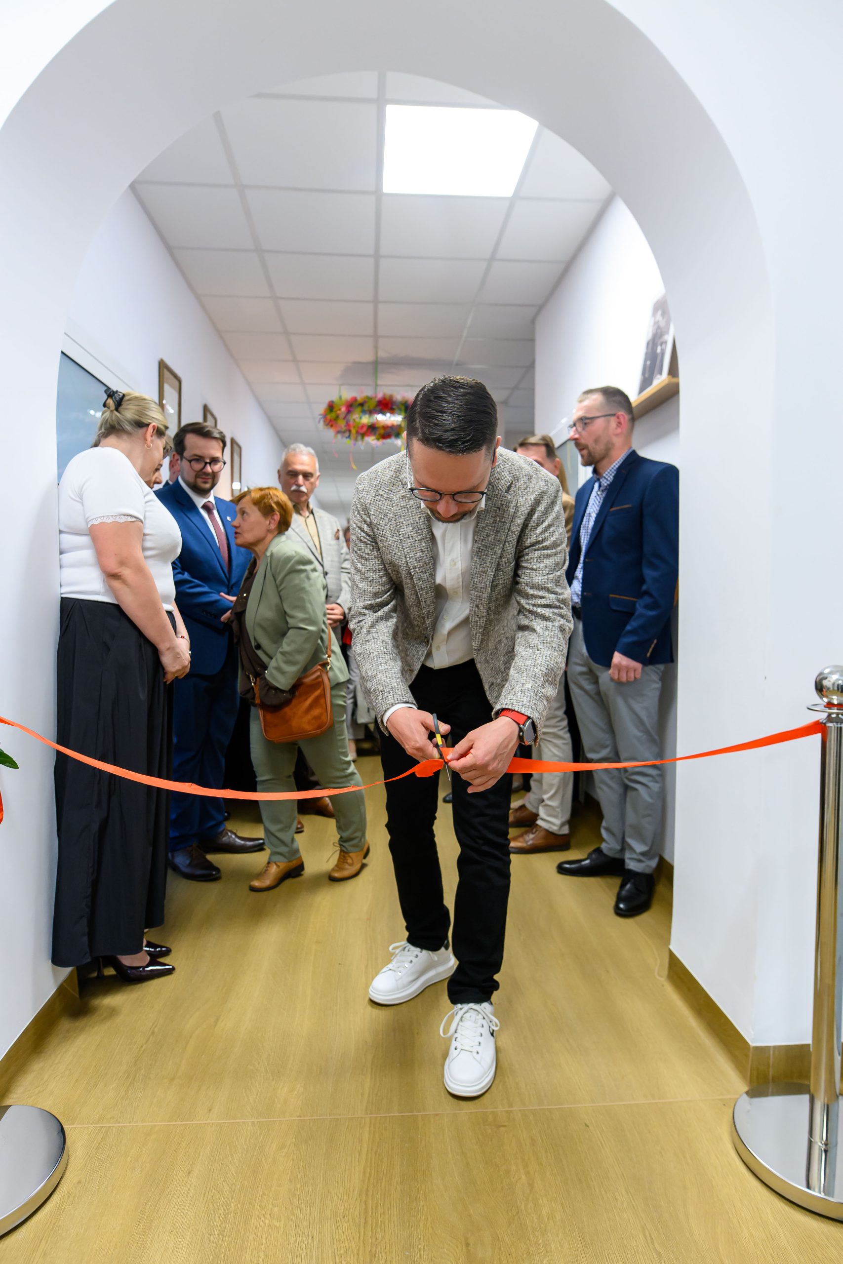 Mężczyzna przecina czerwoną wstęgę w korytarzu z białymi ścianami. Grupa osób w formalnych ubraniach patrzy. Drewniana podłoga.