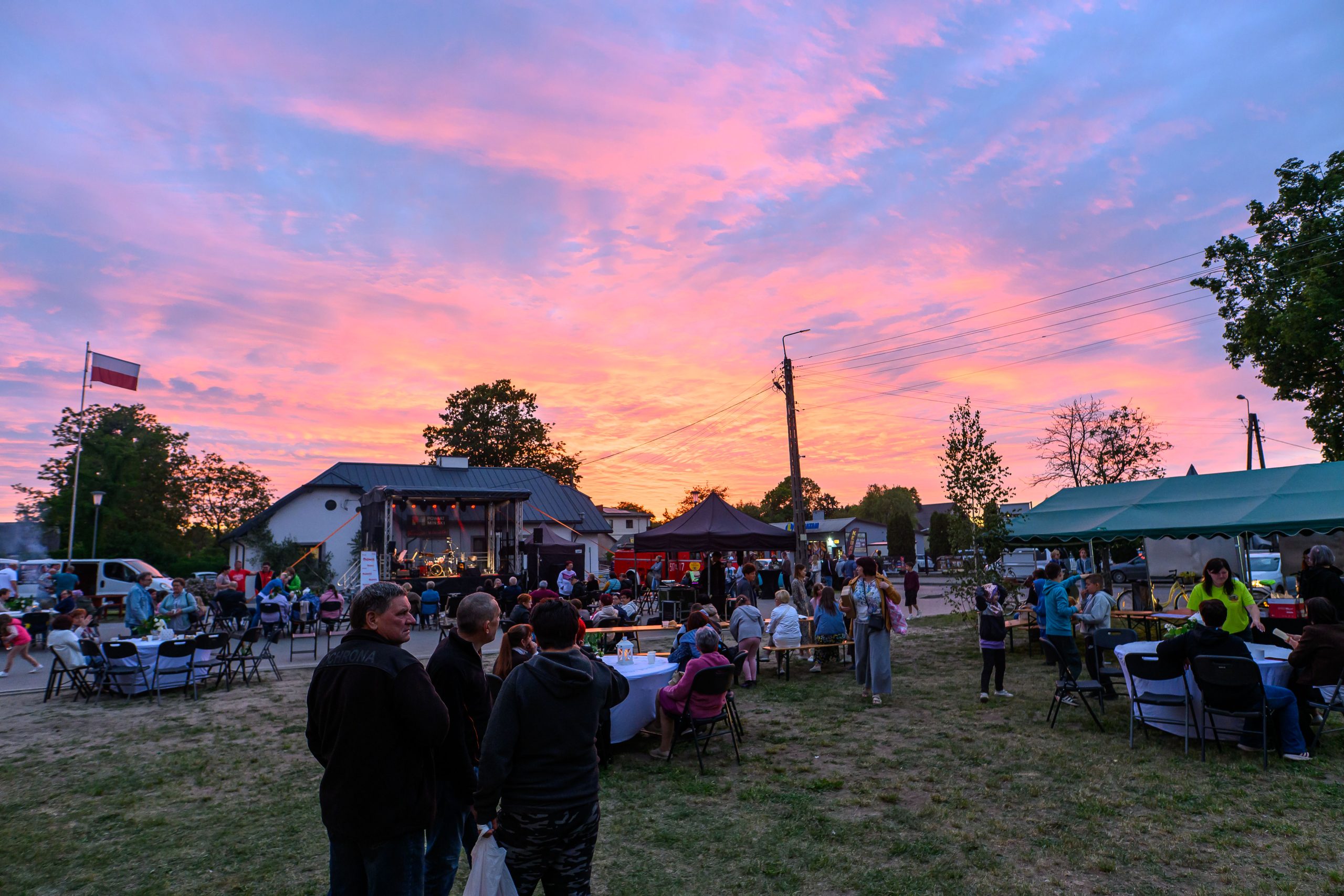 Otwarty festyn z ludźmi na tle różowo-niebieskiego zachodu słońca. Stoiska i scena, wokół zielone drzewa. Atmosfera wieczorna, radosna.