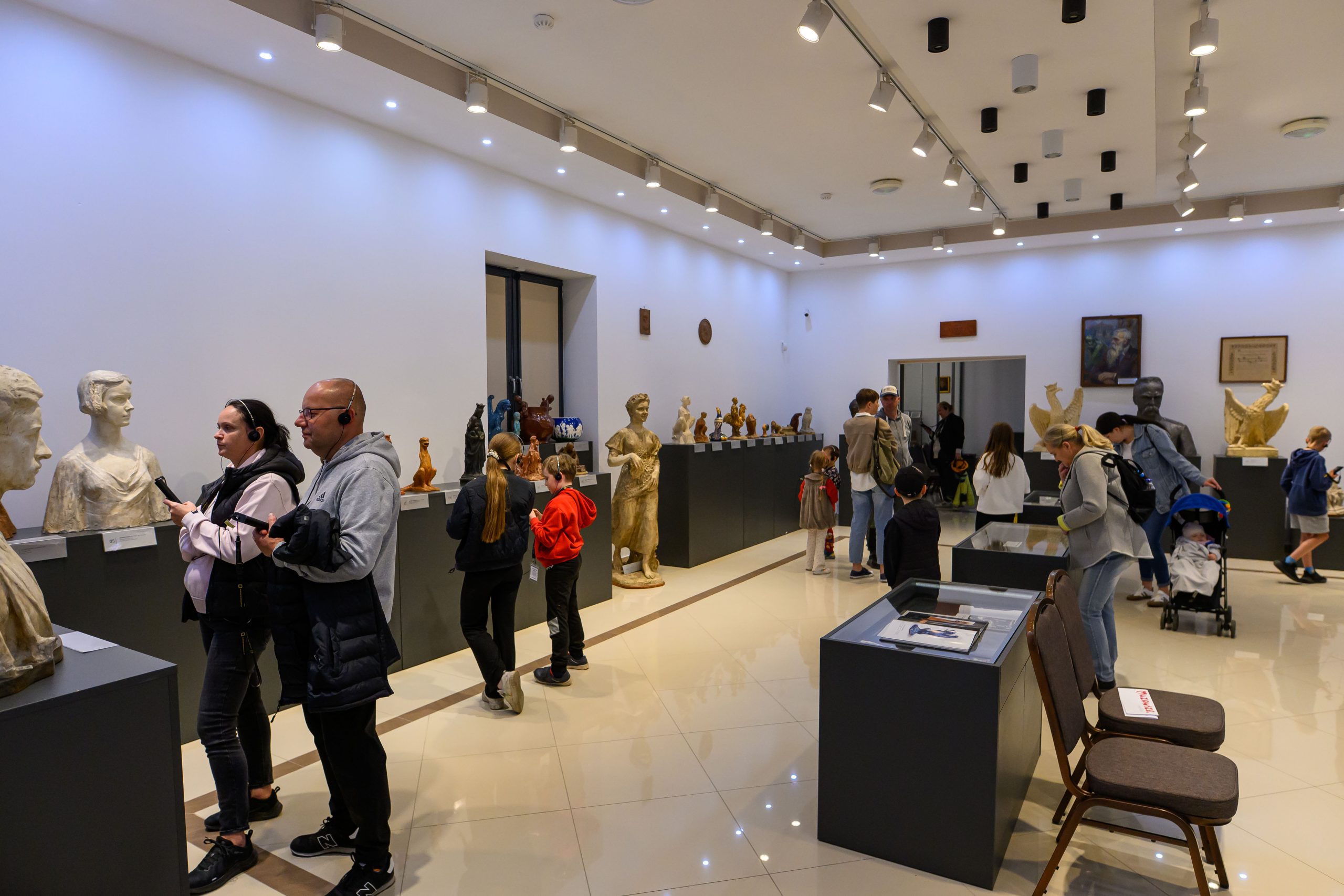 Ludzie zwiedzają galerię pełną rzeźb i popiersi, ściany są białe, podłoga kremowa. Dzieła eksponowane są na ciemnych, prostych cokołach, a sufit oświetlony jest licznymi lampami. W tle w