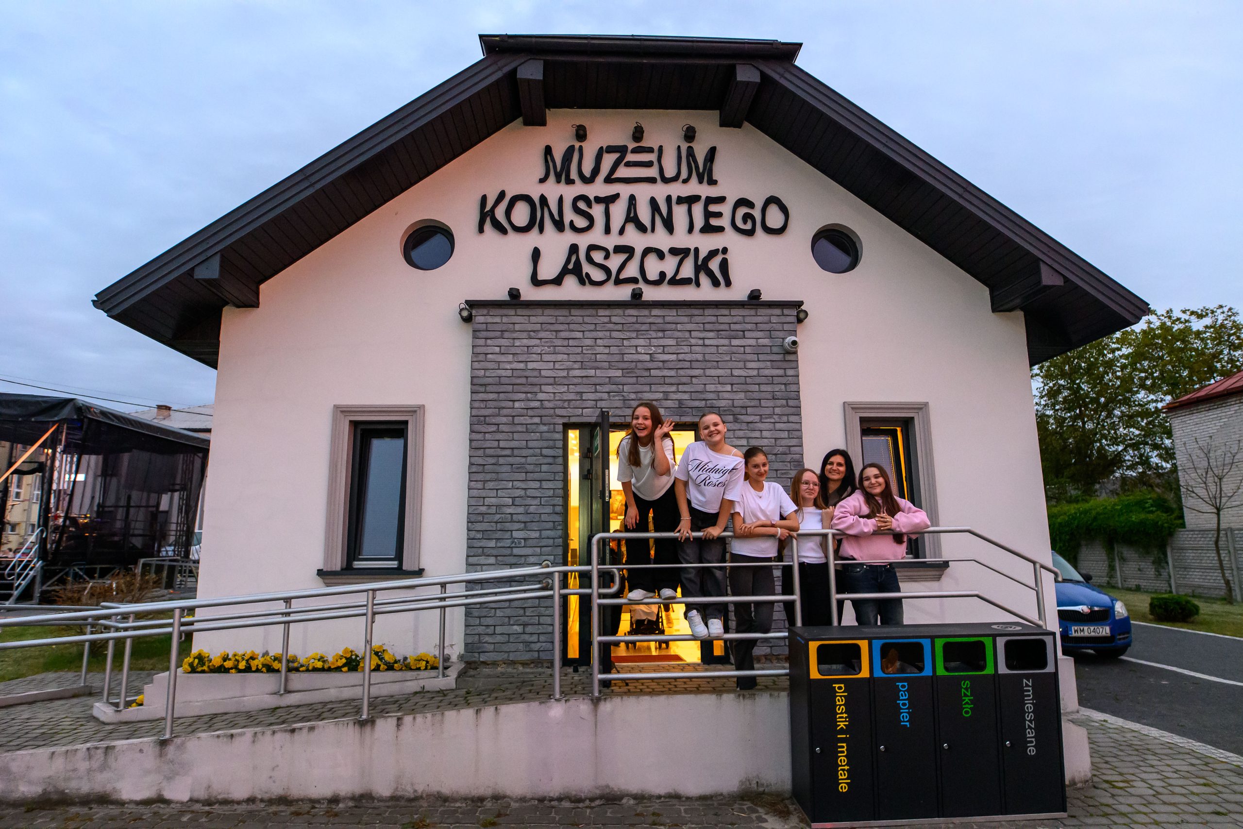 Budynek Muzeum Konstantego Laszczki, o jasnej fasadzie i szarym dachu. Przed wejściem stoi grupa sześciu uśmiechniętych osób. Obok budynku ustawione są kolorowe kosze na śmieci.