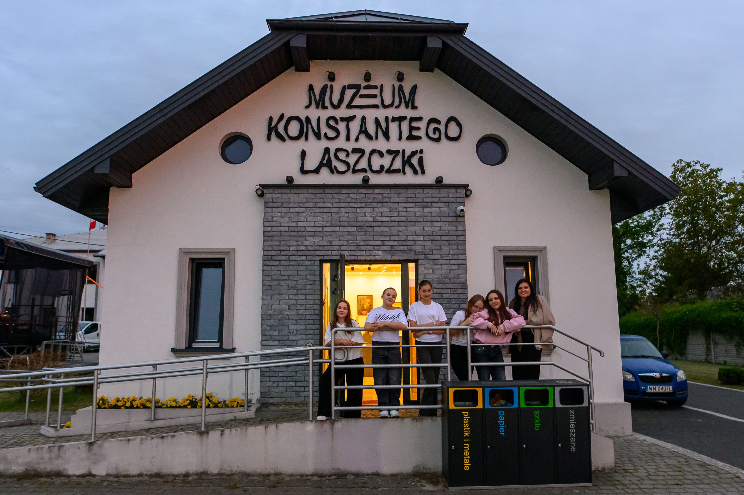 Budynek muzeum w białych i szarych tonacjach z grupą ludzi na tle drzwi. Widoczne kosze na śmieci. W tle wieczorne niebo.