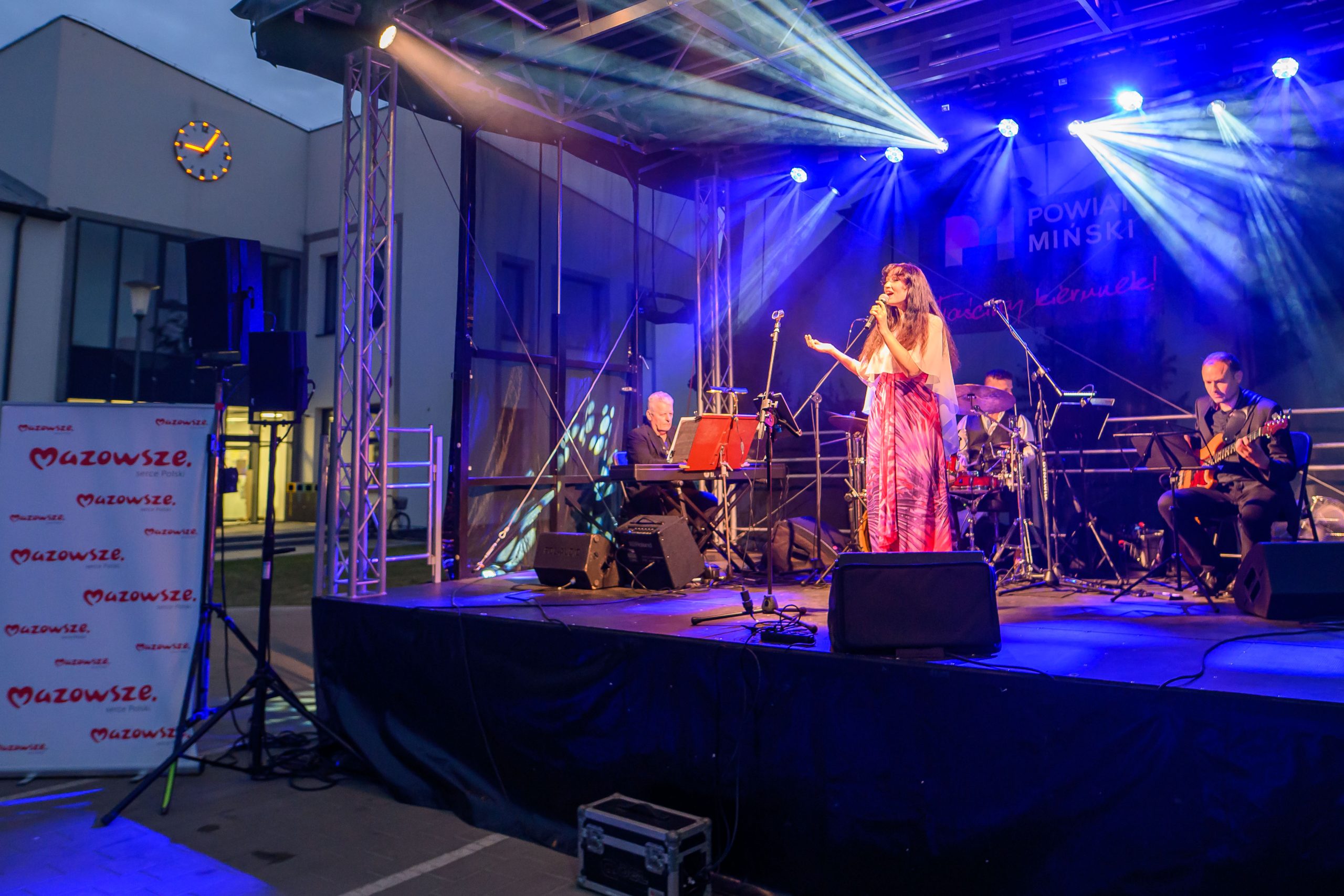 Na scenie w jasnym świetle występuje wokalistka w różowej sukni. Towarzyszy jej zespół muzyczny z bębnami i gitarą. W tle widoczny jest baner z napisem „Mazowsze”.