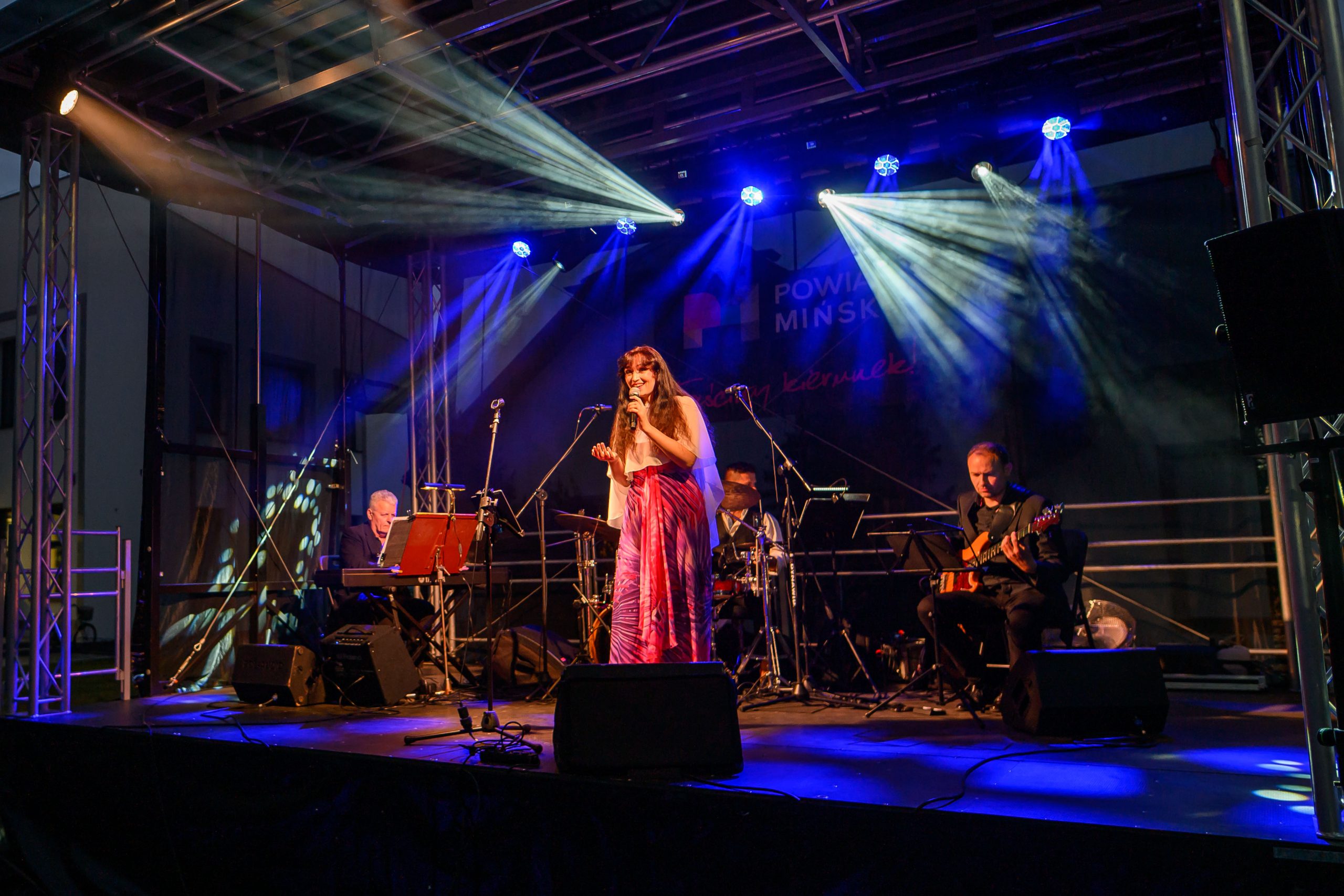 Koncert na scenie; śpiewająca kobieta w różowej sukience, zespół z instrumentami, niebieskie światła w tle.