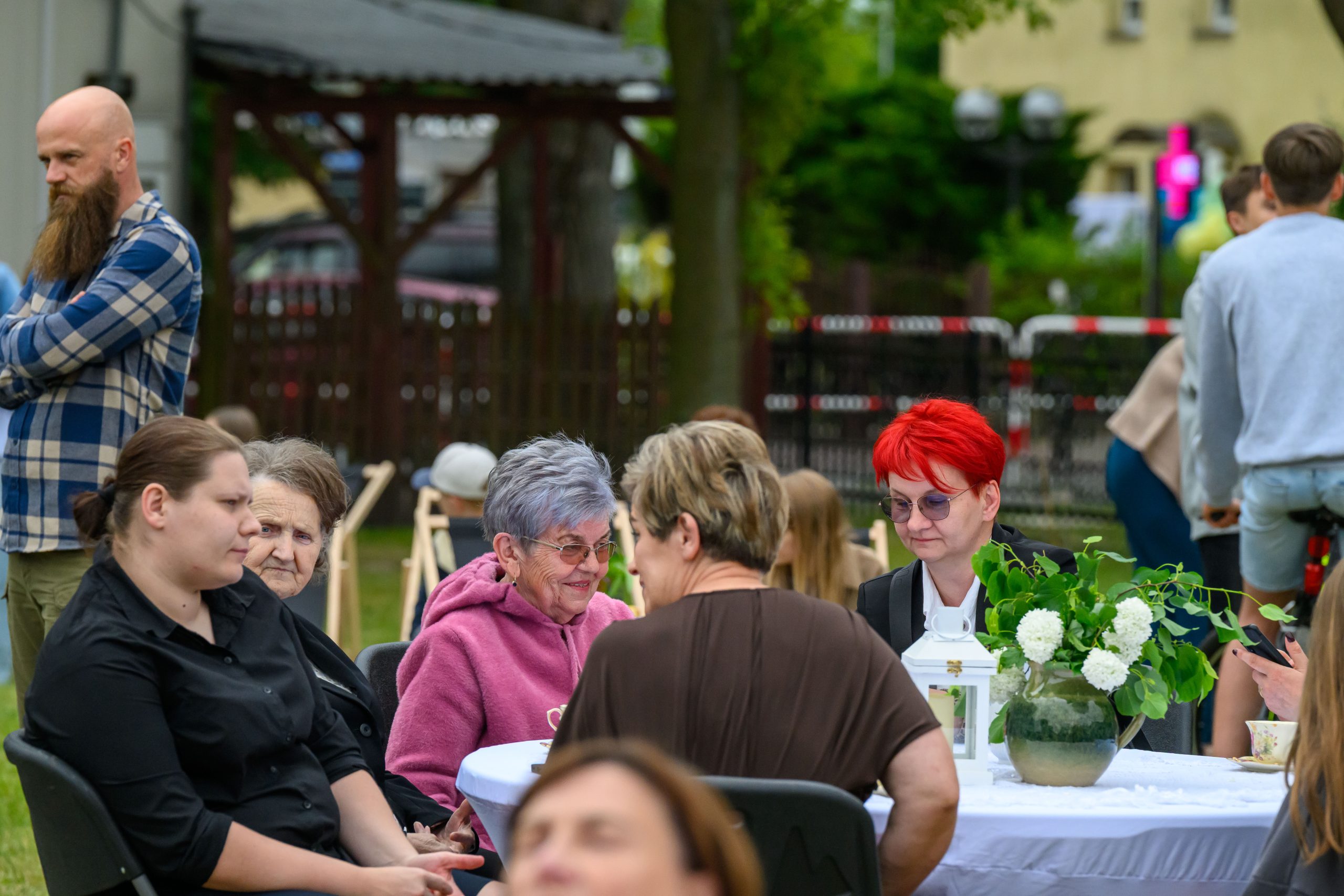 Kilka osób siedzi przy białym stole na zewnątrz, otoczonym zielenią. Na stole jest wazon z kwiatami. W tle widać mężczyznę w koszuli w kratę i zieleni.