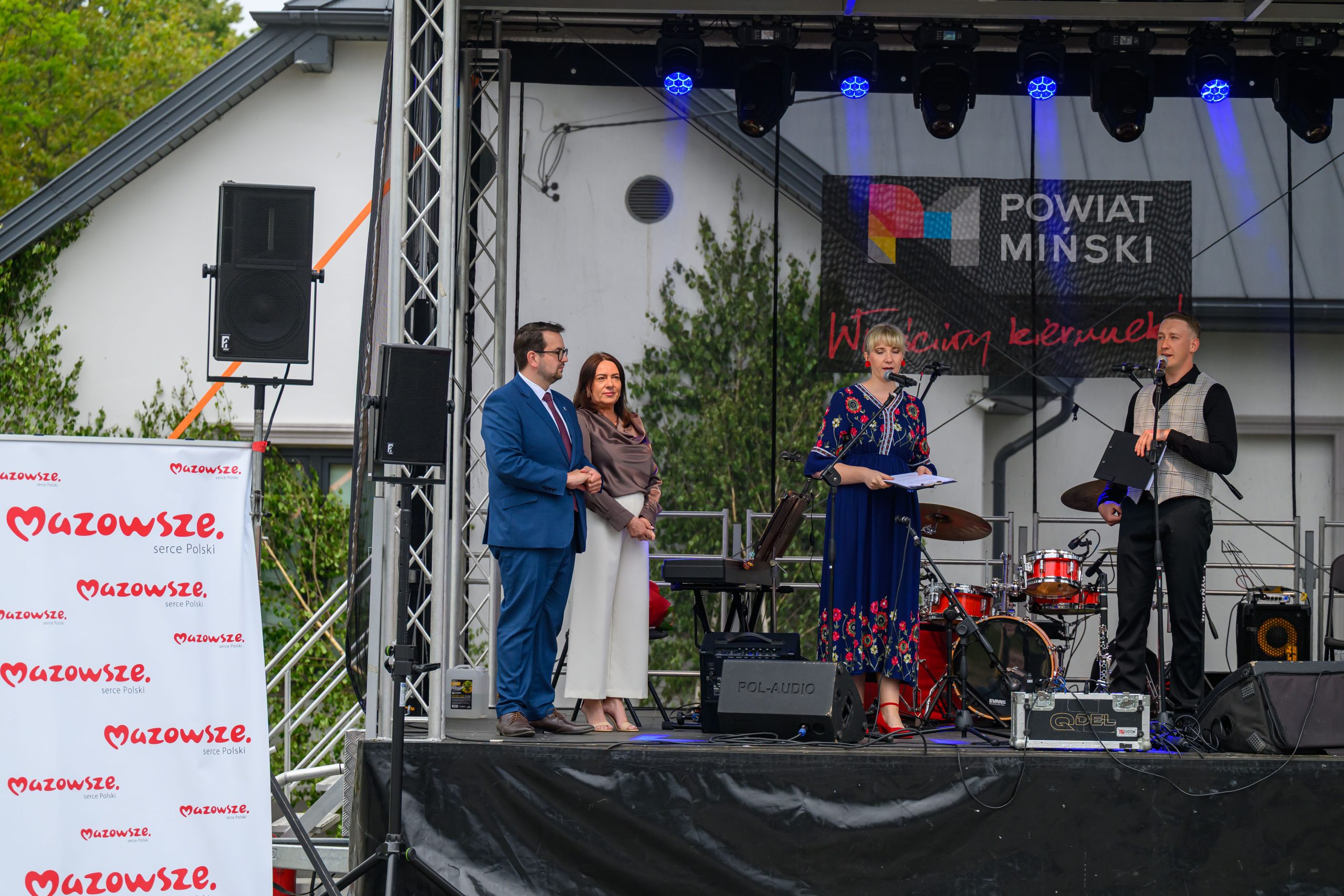 Na scenie przed budynkiem stoją cztery osoby, w tym dwie z mikrofonami. W tle widoczny jest baner Powiat Miński. Na dole widoczny jest napis Mazowsze.