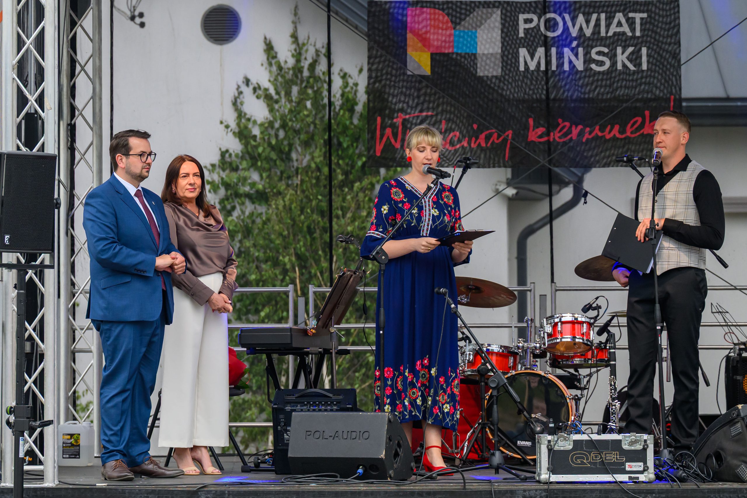Na scenie cztery osoby; kobieta w niebieskiej sukience mówi do mikrofonu. W tle widoczny bęben i zielone krzewy.