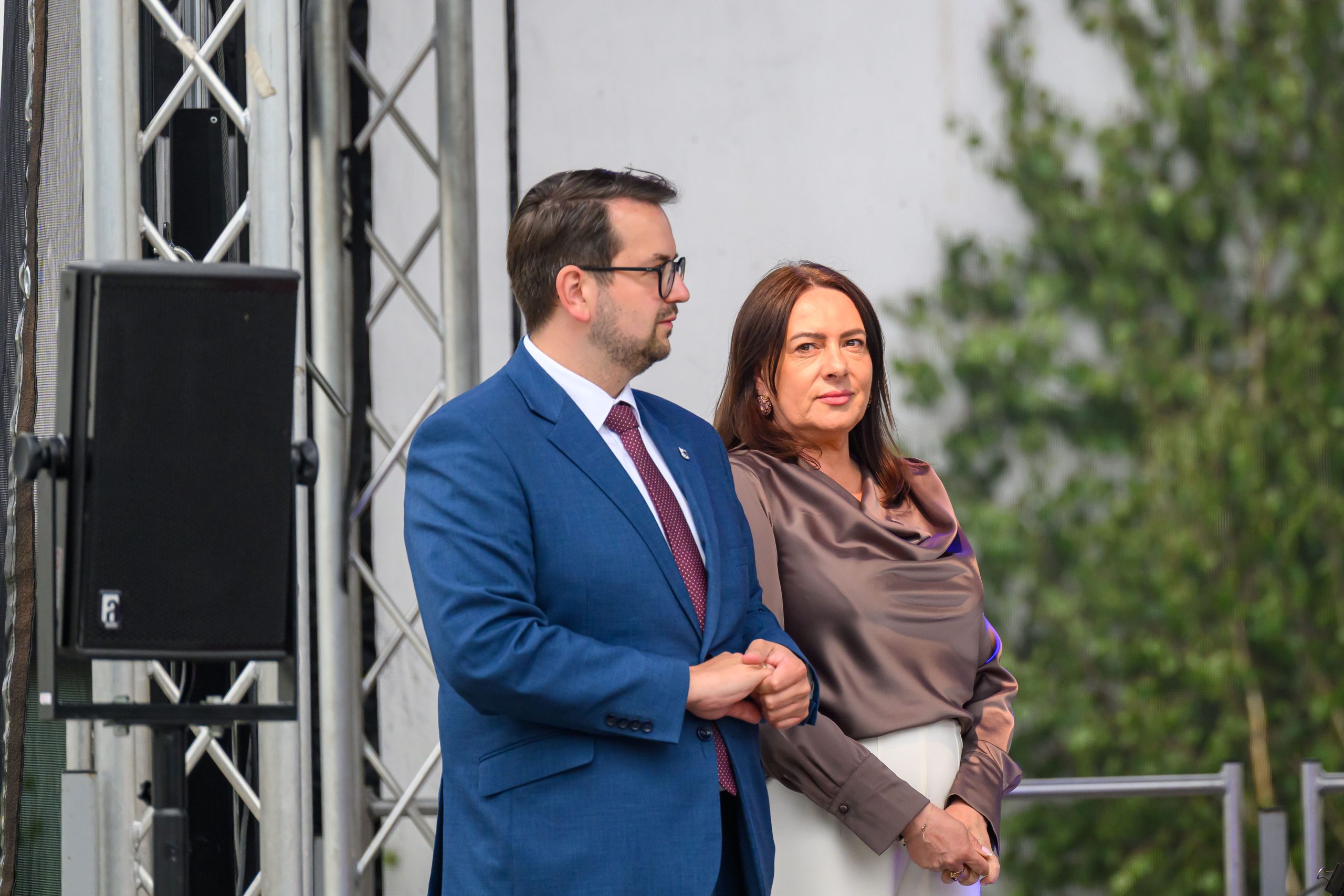 Dwie osoby stoją obok siebie. Mężczyzna w niebieskim garniturze, kobieta w satynowej bluzce, w tle zielone liście. Obok nich widoczna struktura metalowa.