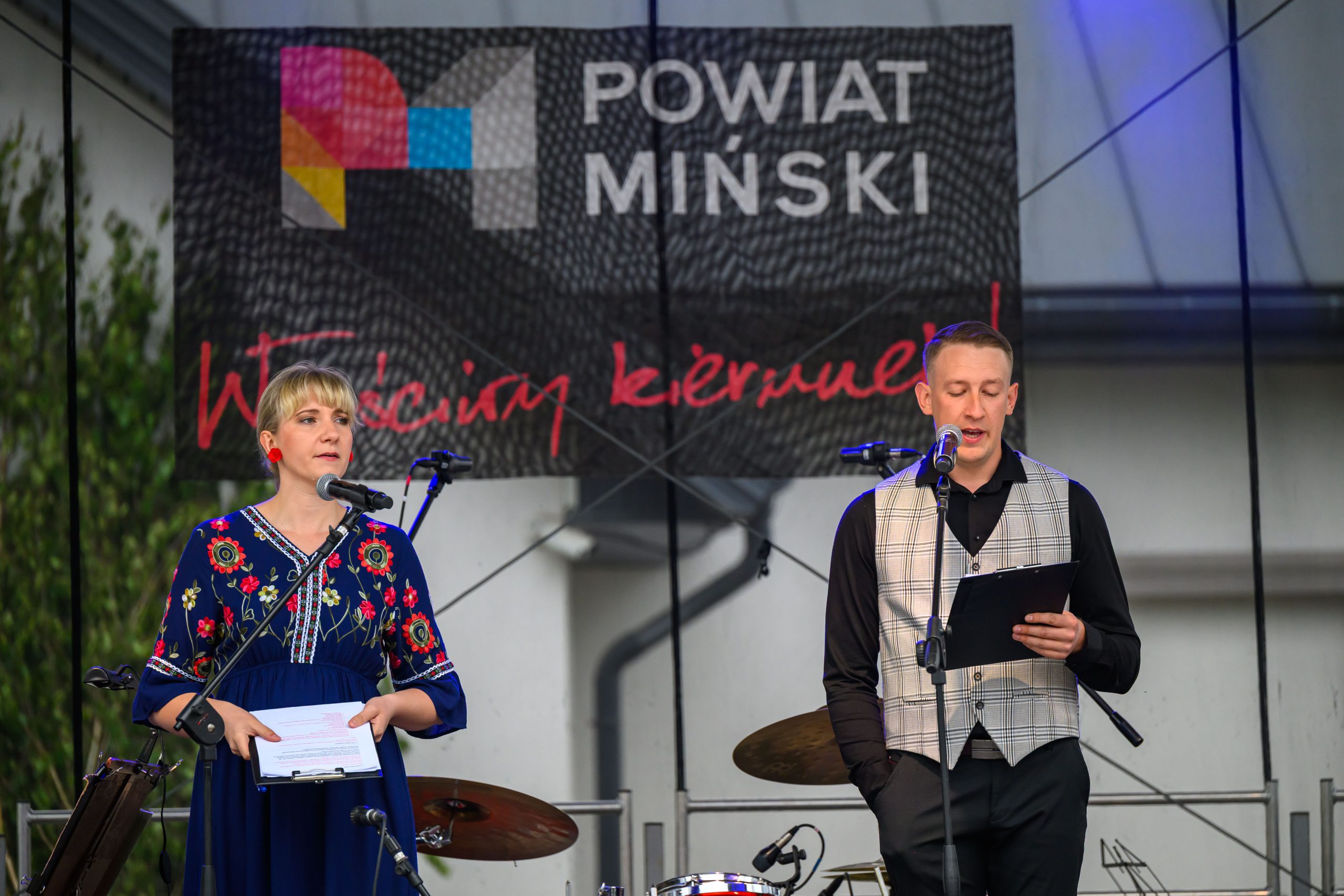Dwie osoby na scenie z mikrofonami; kobieta w niebieskiej sukience, mężczyzna w kamizelce. W tle baner Powiat Miński.