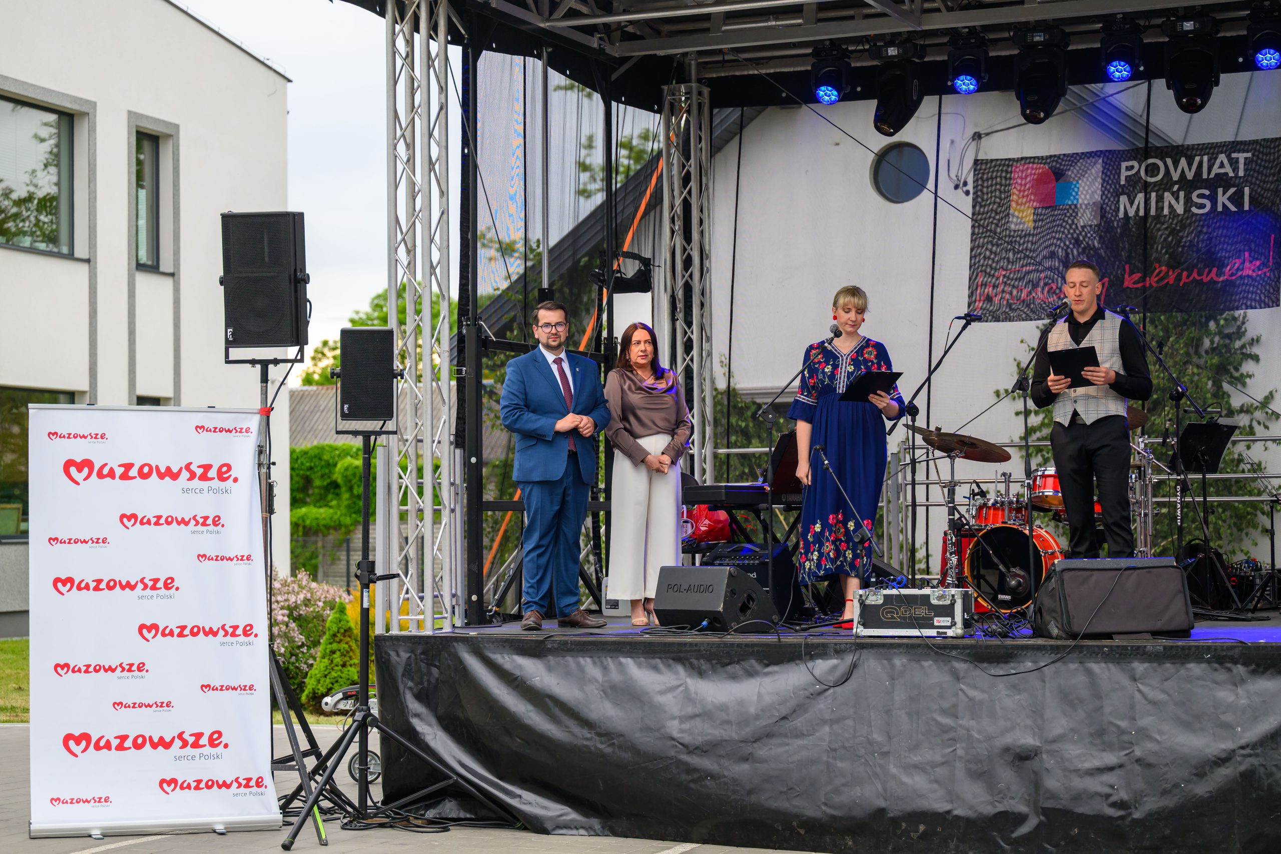 Grupa ludzi stoi na scenie podczas wydarzenia plenerowego. Tło zdobi baner „Powiat Miński”, na scenie są instrumenty muzyczne. Obok sceny stoją głośniki i biały roll-up „Mazowsze”.