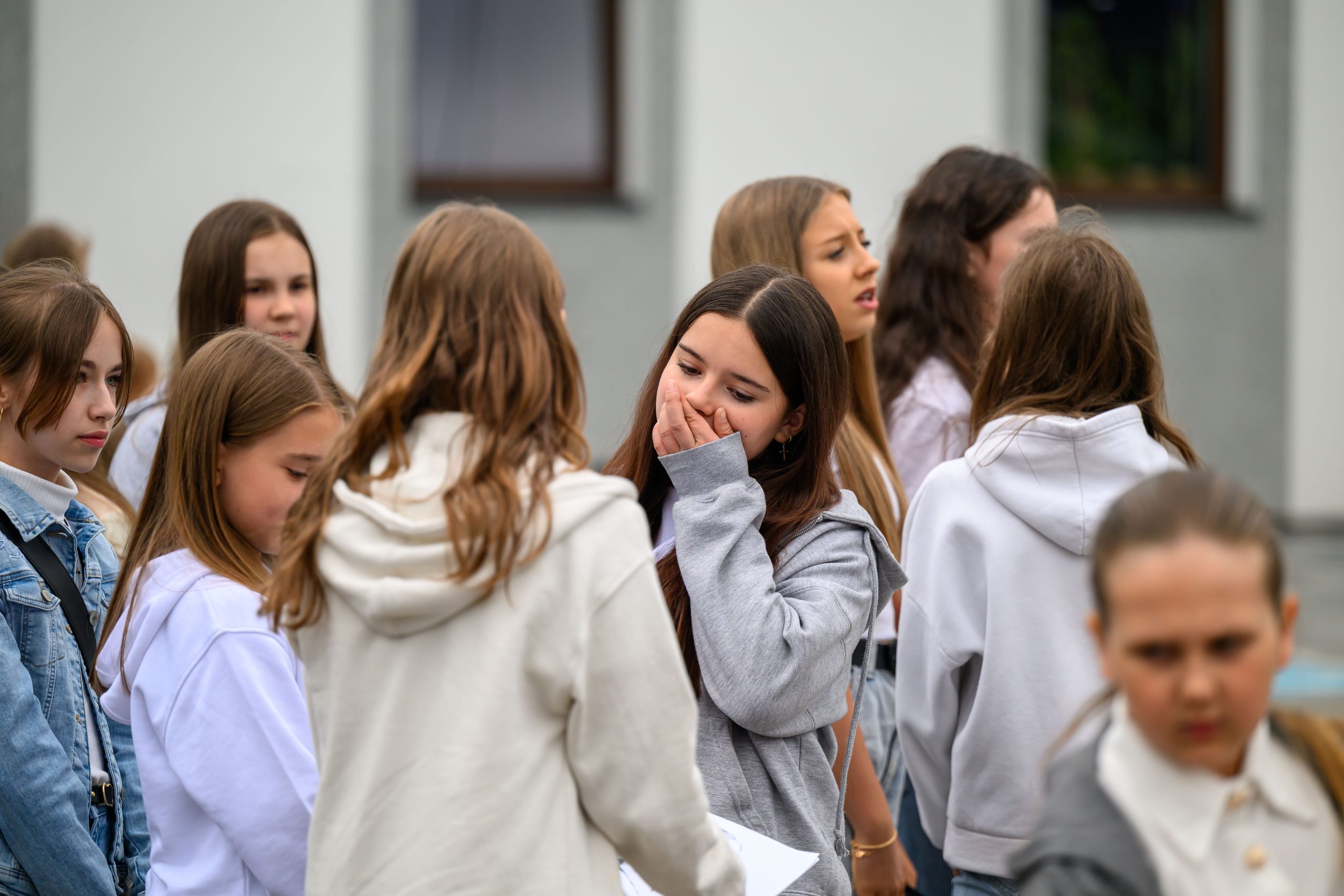 Grupa dziewcząt w jasnych bluzach stoi na zewnątrz, przed szarym budynkiem. Kilka osób rozmawia i śmieje się. Tło jest rozmyte, skupiając uwagę na postaciach.