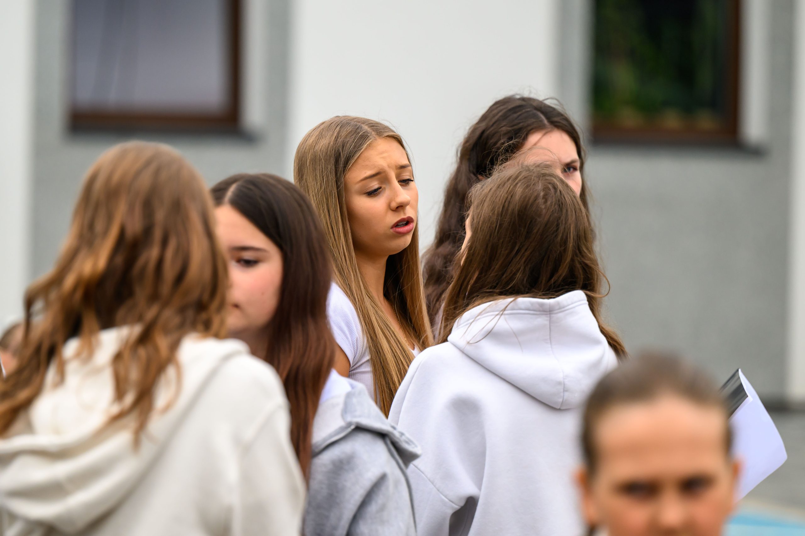 Grupa młodych osób na zewnątrz budynku, w jasnych ubraniach. Dominują odcienie brązu i bieli. W tle szare ściany.