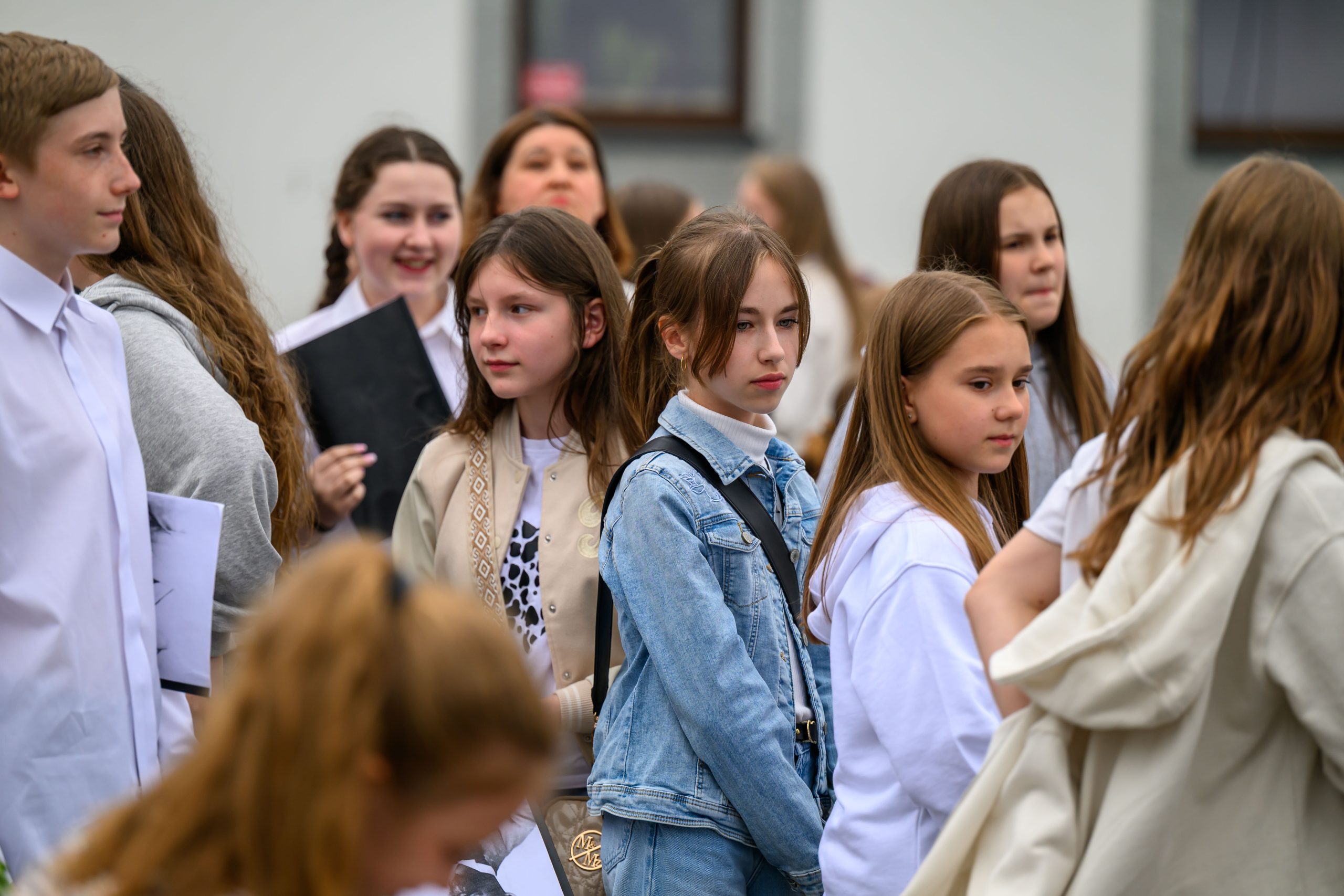 Grupa młodzieży w jasnych ubraniach stoi na zewnątrz. Dominuje kolor biały i szary. Tło jest neutralne, skupione na postaciach.