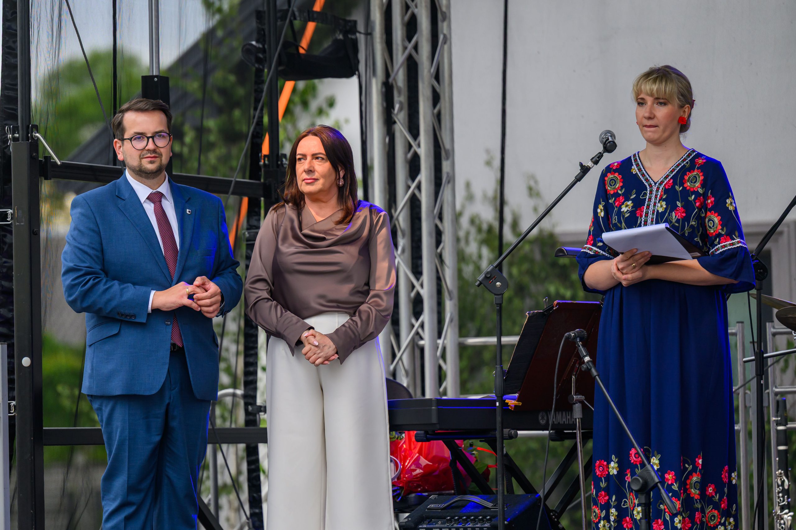 Na zdjęciu widać trzy osoby na scenie: dwie stojące obok siebie i jedną przy mikrofonie. Pierwsza osoba w niebieskim garniturze, druga w ciemnej bluzce i białych spodniach, trzecia w niebieskiej sukni z kwiatami.