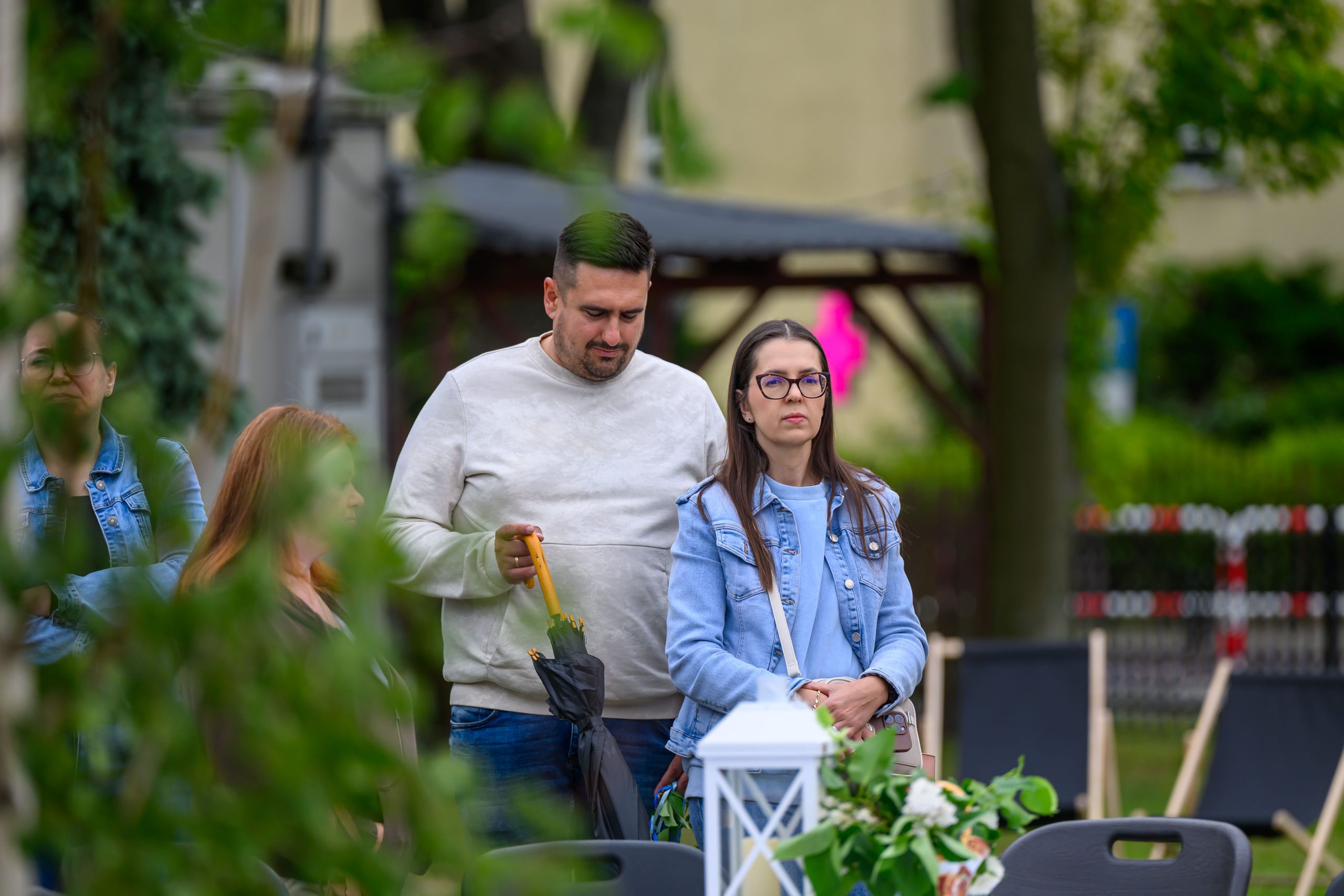 Zdjęcie przedstawia mężczyznę i kobietę na zewnątrz, z zielenią w tle. Mężczyzna trzyma parasol, oboje są w casualowych ubraniach. Kobieta ma ciemne okulary i jasną kurtkę jeansową.