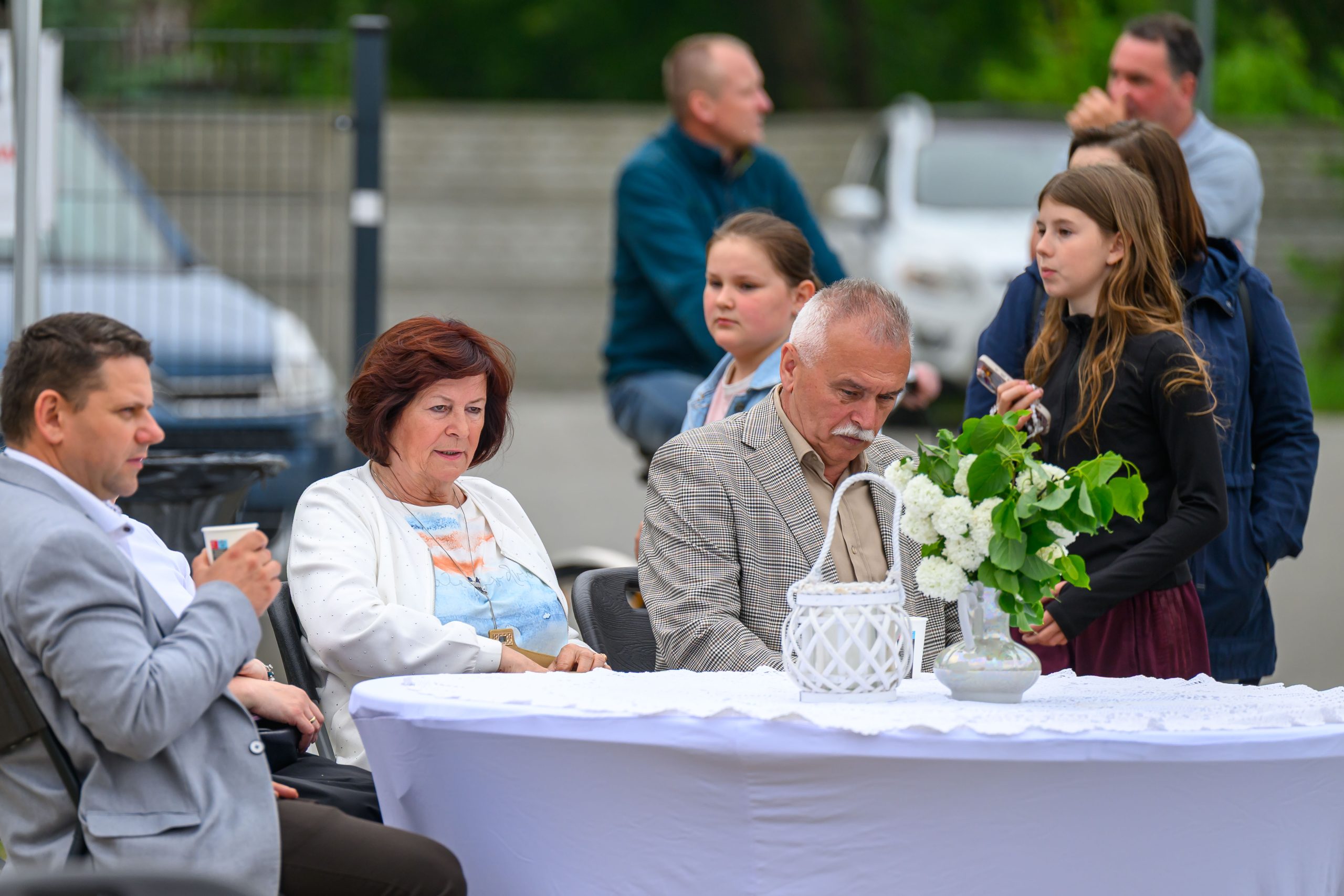 Na zdjęciu widać ludzi przy stole z białym obrusem i zielonym bukietem. Tło tworzą niebieskie i szare detale zewnętrzne. Każda osoba przyjmuje różne pozy i zajęcia.