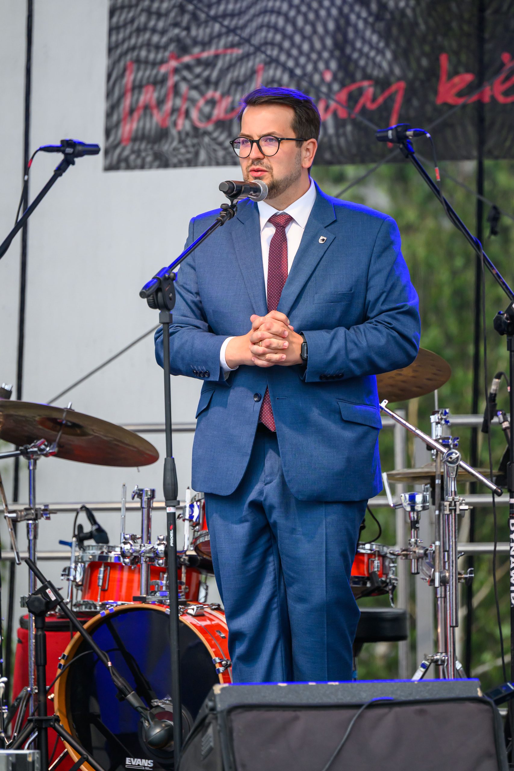 Mężczyzna w niebieskim garniturze przemawia na scenie. Za nim znajduje się zestaw perkusyjny i transparent z napisem. Scena oświetlona, w tle drzewa.