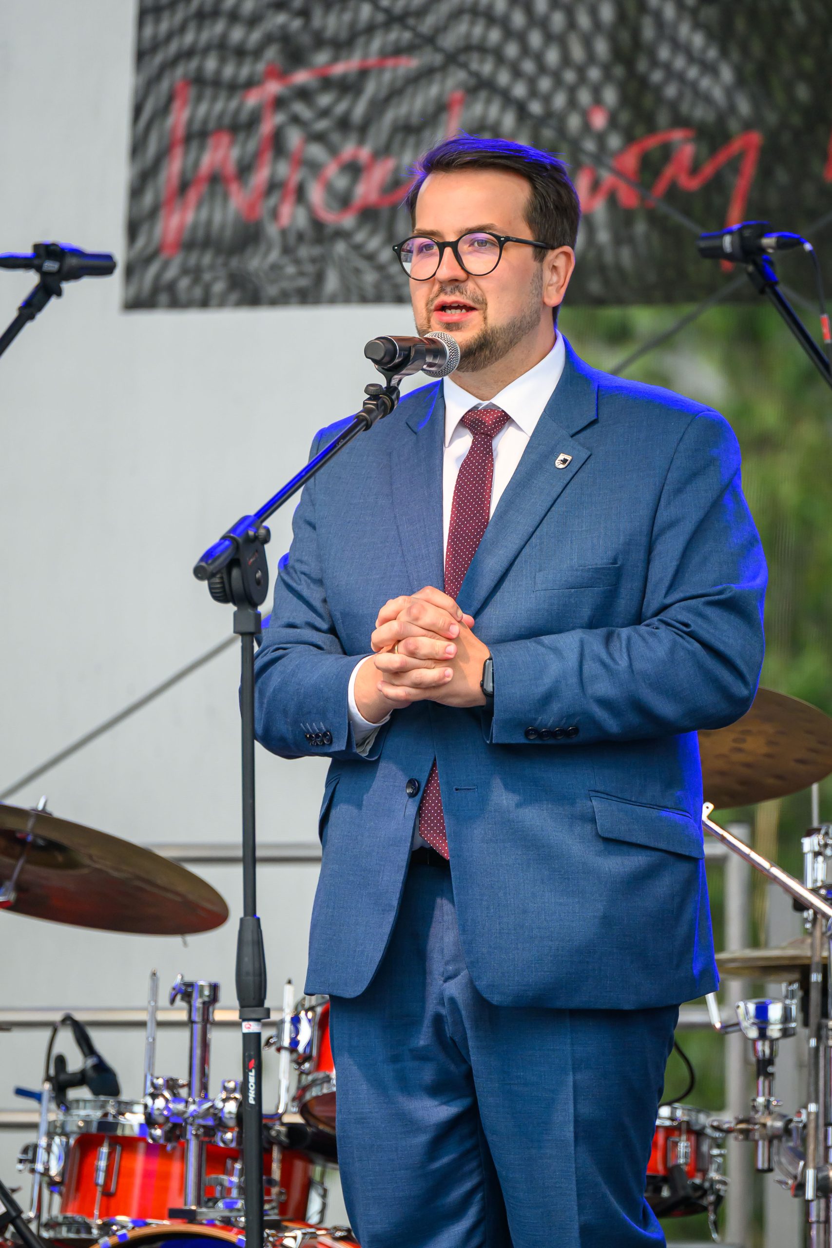 Mężczyzna w niebieskim garniturze przemawia na scenie. W tle znajdują się perkusja i napis w czerwieni. Scena jest oświetlona światłem dziennym.