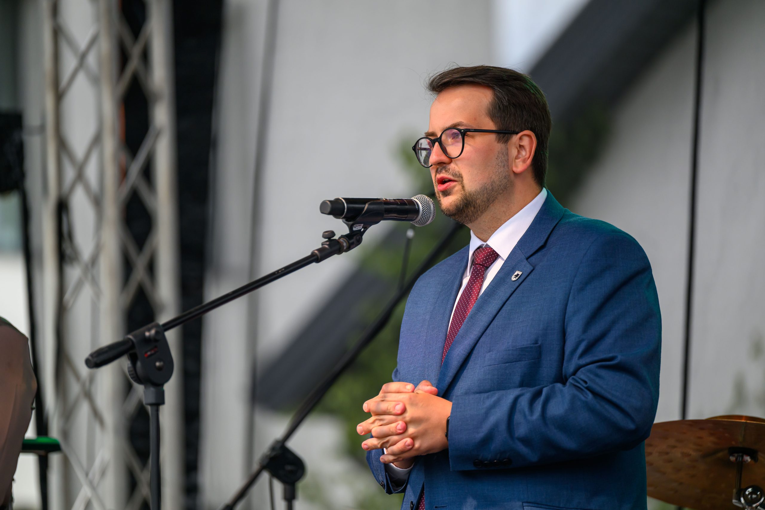 Mężczyzna w niebieskim garniturze przemawia do mikrofonu. Scena na świeżym powietrzu, w tle metalowe rusztowanie. Wygląda formalnie.