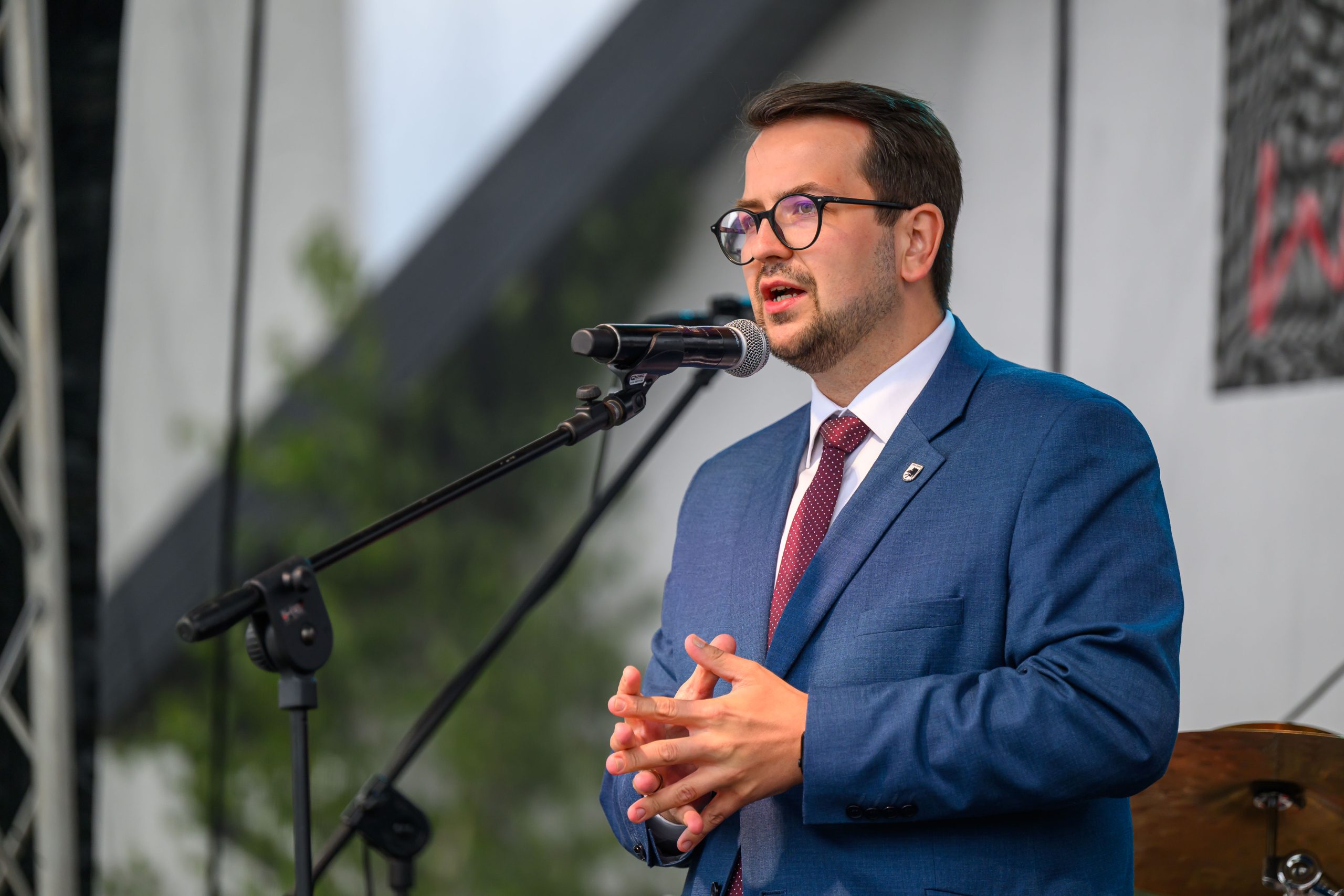 Mężczyzna w niebieskim garniturze przemawia do mikrofonu. Ma okulary i ciemne włosy. Tło jest rozmyte, z zielenią i szarościami.