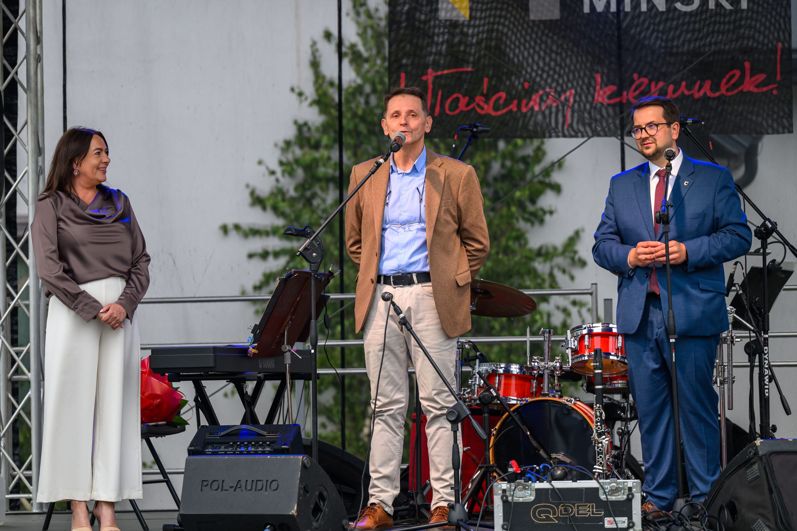 Na scenie stoi trzech ludzi z mikrofonami. W tle widać sprzęt muzyczny i zielone liście. Mężczyzna w środku mówi do mikrofonu.