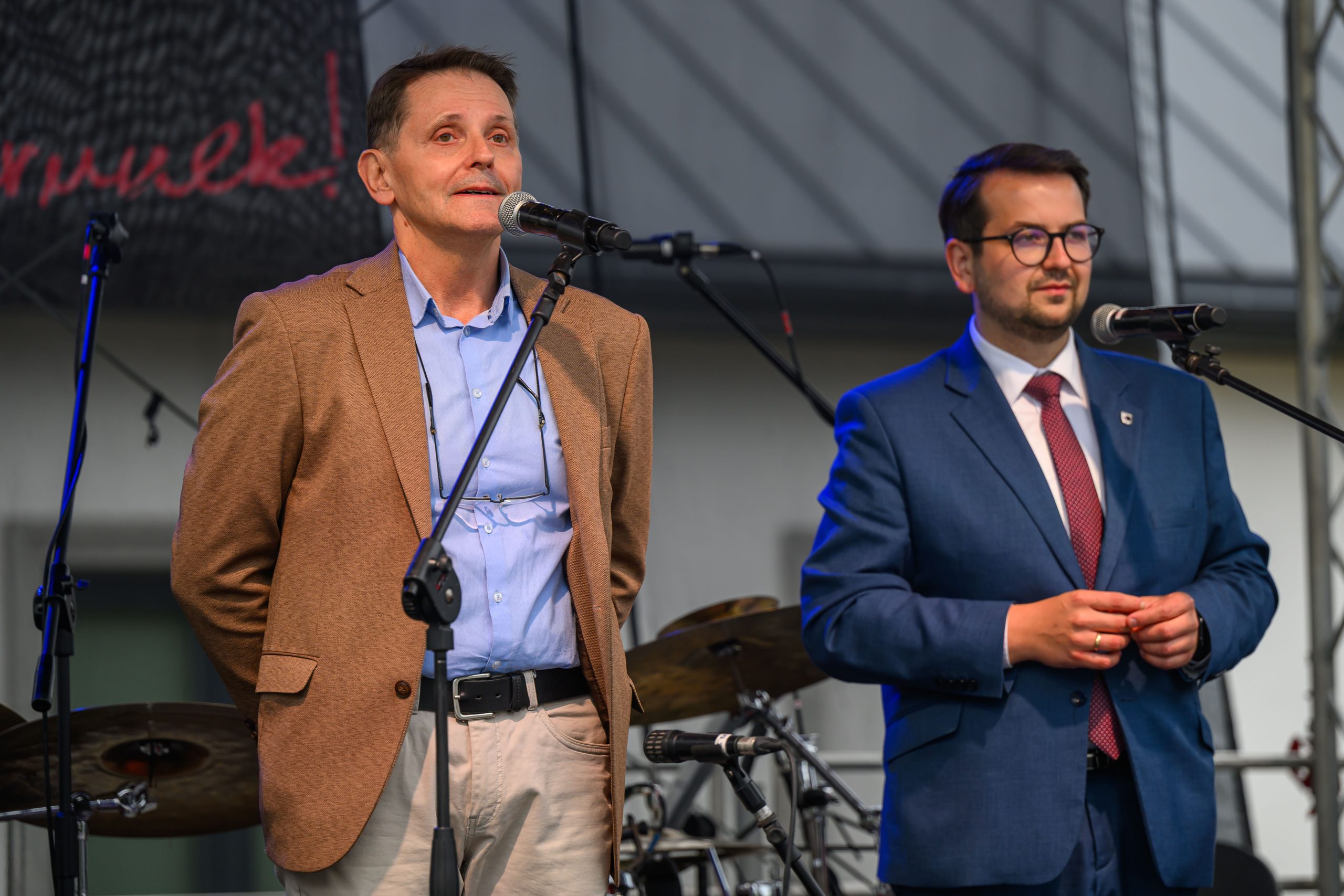 Dwóch mężczyzn stoi przed mikrofonami na scenie. Jeden ma brązową marynarkę, drugi niebieski garnitur. W tle widać instrumenty muzyczne.