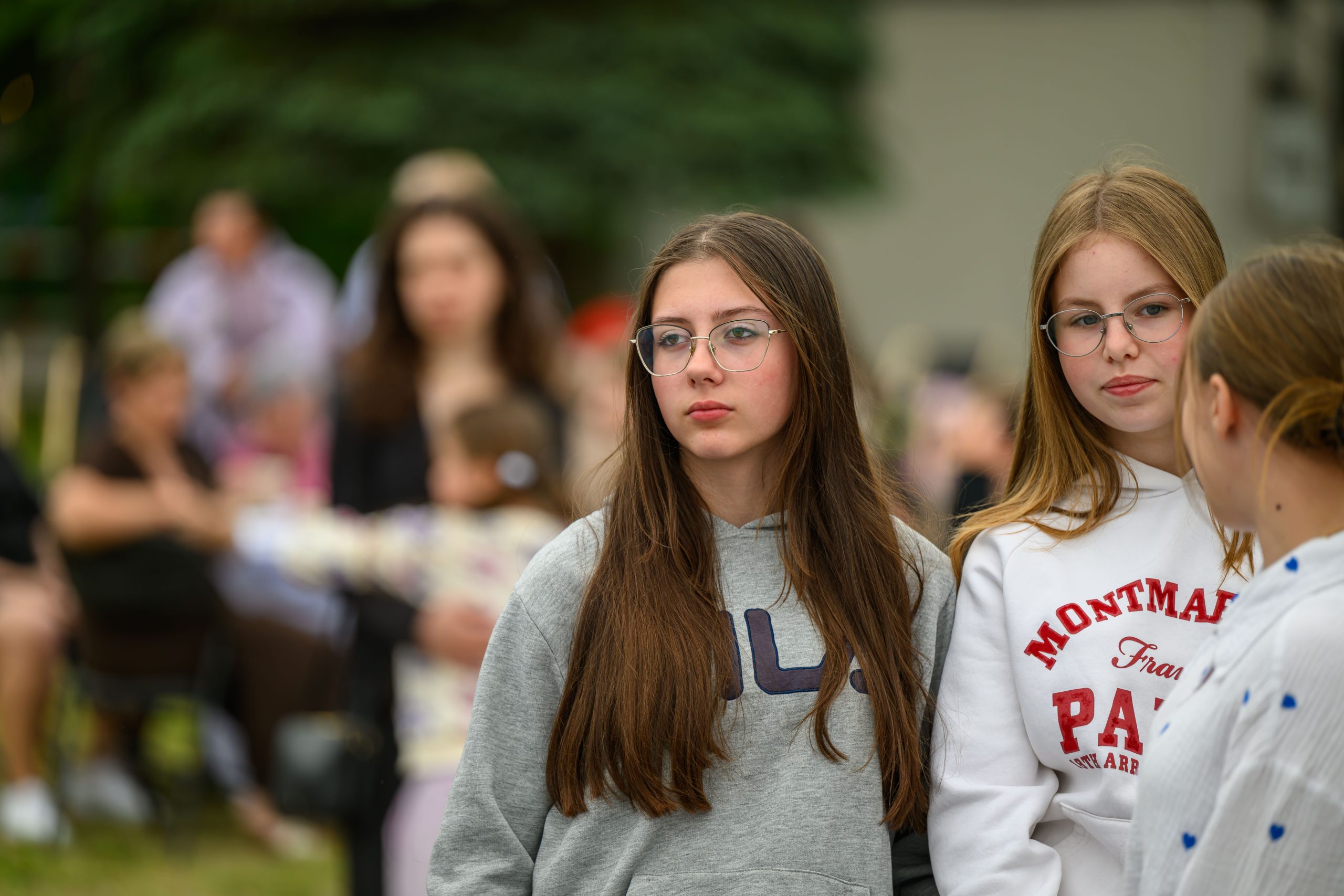 Grupa młodych osób stoi na zewnątrz. Ubrani są w sportowe bluzy, w tle widać rozmazane postacie. Tło jest zielone, nadając wrażenie parku.